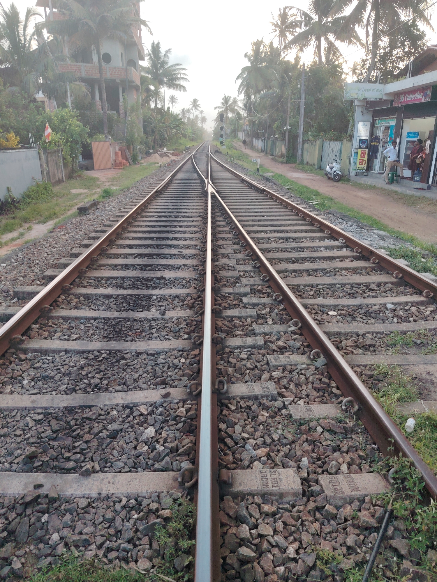 The image shows a set of railroad tracks in an outdoor environment. The tracks are made of steel rails supported by concrete sleepers, which are embedded in a bed of gravel. The tracks converge in the distance, suggesting a junction or a switch. On the left side of the tracks, there is an unfinished building with exposed brickwork and palm trees. A small white and red flag is visible on a fence. On the right side of the tracks, there are several shops with awnings and signs. A white scooter is parked near the shops. Two people are standing outside one of the shops. The sky is bright and hazy, indicating daylight. The overall scene depicts a railway line in what appears to be a developing or semi-urban area, possibly in a tropical region due to the presence of palm trees.