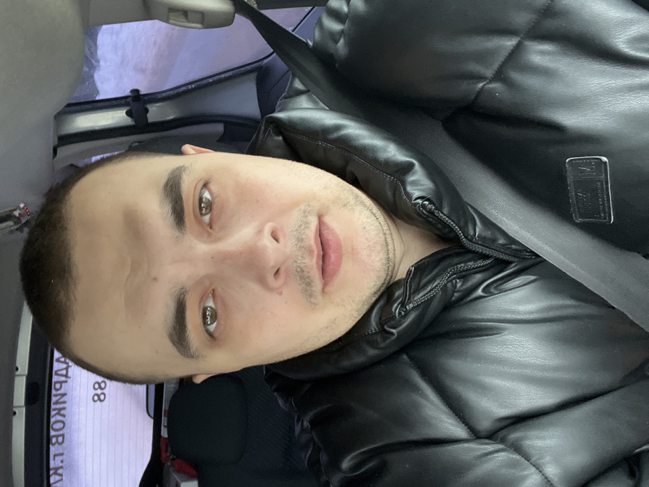 A male individual is depicted in a close-up, front-facing portrait, seated inside a vehicle. He possesses short, dark hair, brown eyes, and light stubble on his chin and upper lip. He is wearing a black, quilted, high-collar jacket, and a gray seatbelt is secured across his chest. In the background, visible through the vehicle's rear window, is reversed Cyrillic text and the number "88" adhered to the glass. To the right, an indistinct, blurry outdoor environment, possibly snowy or overcast, is visible through a side window. A phone mount or similar accessory is attached to the vehicle's interior pillar on the right. The image provides no specific visual cues to determine the exact location within the city of Rogachovo, Russia.
