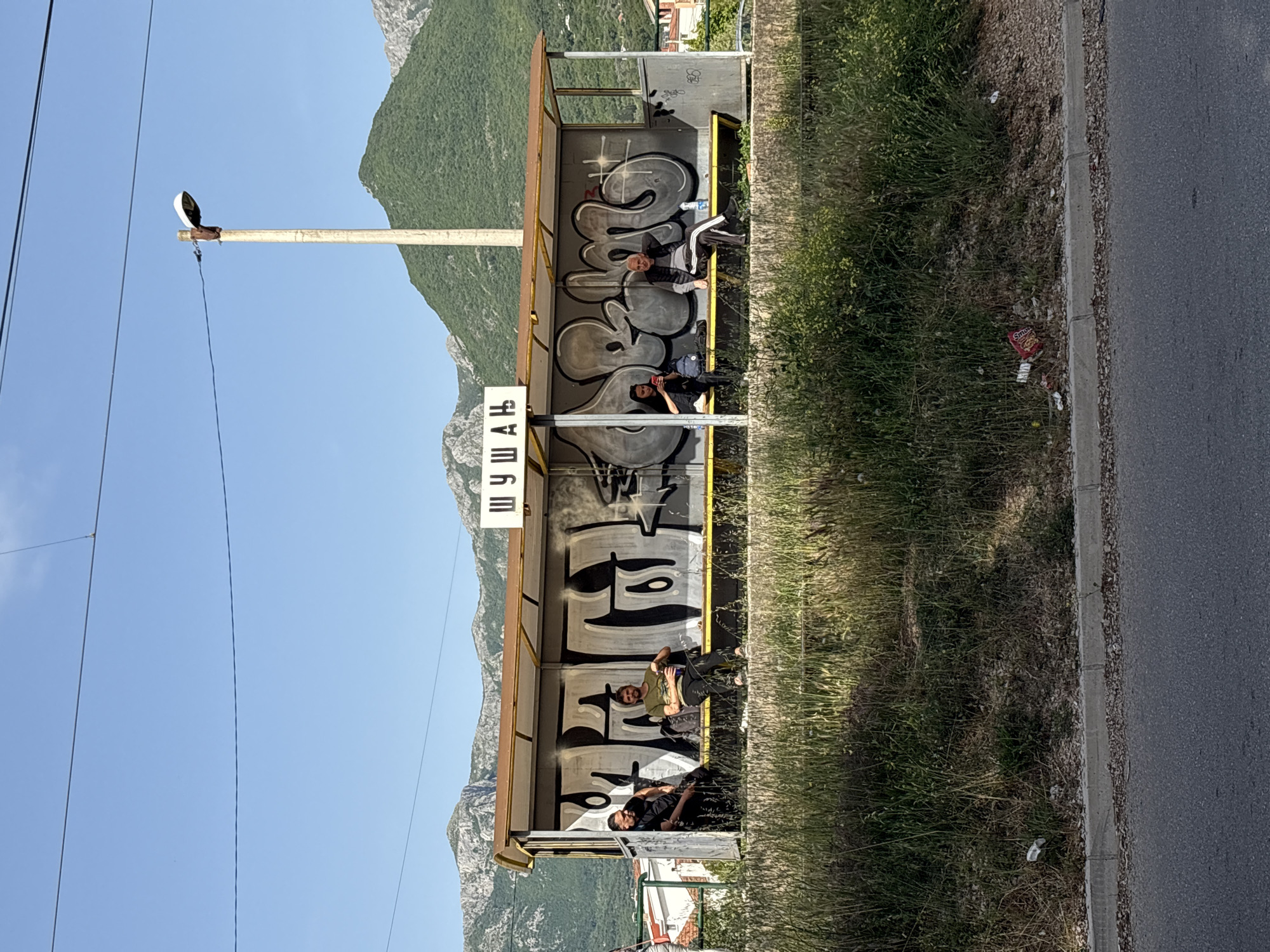 The image depicts a bus stop located in Bar, Montenegro. The bus stop is constructed of metal with a transparent roof and is covered in graffiti. The name of the bus stop is written in Cyrillic script.
Four individuals are sitting inside the bus stop. The person closest to the camera is lying down, another is on their phone. Further back another is on their phone. The last person is laying down.
Outside the bus stop, there is a green patch of grass, a street light on a pole, and a road. In the background, there is a mountain range under a clear blue sky.