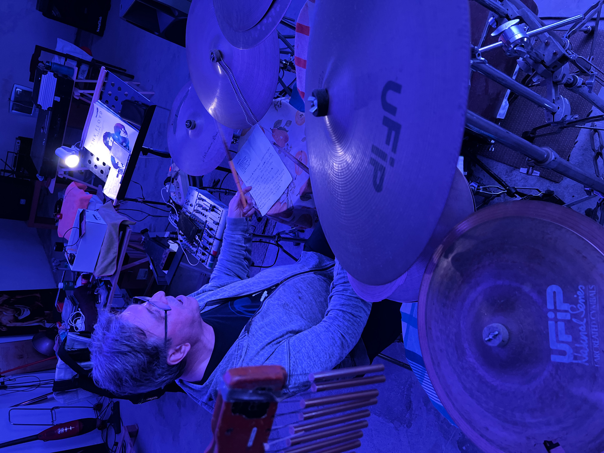 The image depicts an adult male engaged with a drum kit in an indoor music studio, understood to be located in Santeramo in Colle, Italy. The scene is bathed in a predominant blue ambient light, with a singular bright white light illuminating a music stand.
The man, appearing middle-aged with greying hair, wears glasses, a dark t-shirt, and a grey hooded zip-up sweatshirt. He is seated on a drum stool, looking towards the right side of the frame with his mouth slightly ajar, as if speaking or singing. His right hand holds a drumstick, poised above a cymbal. To his immediate left is a small audio mixer with numerous knobs and faders. A handwritten piece of paper, likely music notes or lyrics, rests on a table covered with a white and orange patterned tablecloth, situated between the man and the drum kit.
The drum kit features several cymbals; one in the foreground is clearly branded "UFIP National Series Arc Created Cymbals," and another visible to the right is marked "Medium Thin." To the far left, hanging next to the man, is a set of golden-colored chimes suspended from a red bar.
In the mid-ground, a black music stand holds a book or sheet music titled "PINK FLOYD," displaying album artwork (likely *Wish You Were Here*). The focused white light source is clipped to this stand.
The background on the left reveals a red stick vacuum cleaner leaning against a wall, alongside other assorted objects. Above a table, stacked items resemble folded clothing or blankets, and a large white box is visible. A dark, framed image or poster of a classical figure adorns the far left wall. On the right side of the background, various tables hold electronic equipment, including a classic CRT monitor and large black speakers, one bearing the "SoundTech" brand. A blue water bottle rests on one of these tables. The floor is concrete, partially covered by small dark rugs or mats beneath the drum kit. The overall environment is a functional, somewhat cluttered space dedicated to musical practice.