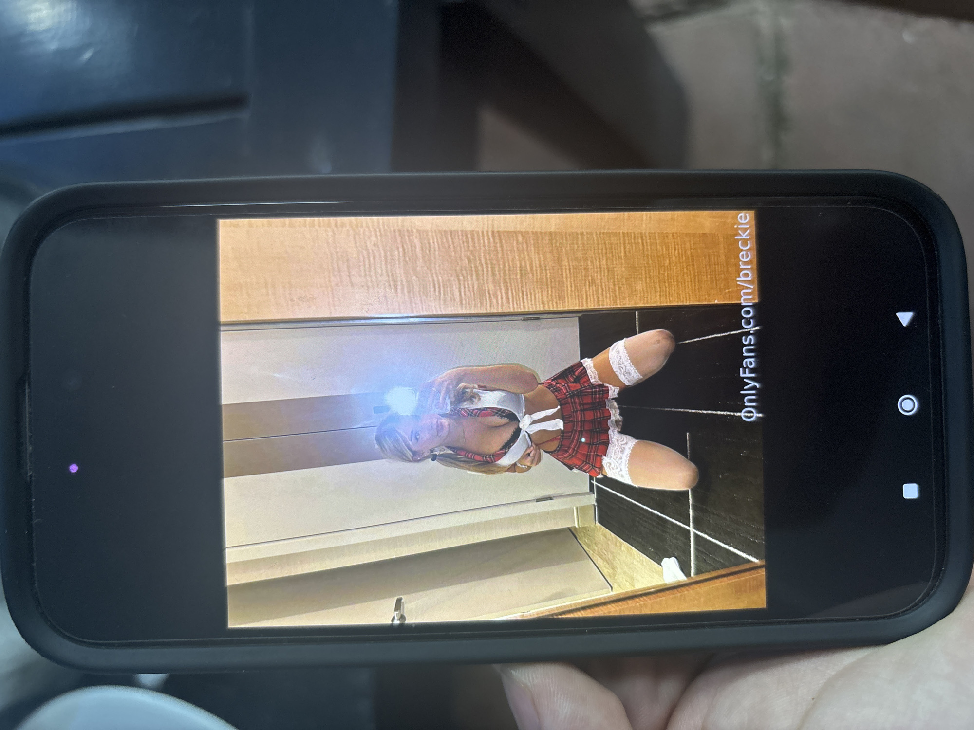 A dark-colored smartphone, held by a person's left hand, displays an image of a woman. The woman, with blonde hair styled in pigtails, is kneeling on a dark floor with light-colored lines, taking a mirror selfie with her own light-colored smartphone, which emits a bright flash. She is dressed in a schoolgirl-style costume, featuring a white tie-front crop top over a visible black bra, a red plaid mini-skirt with white lace trim, and white lace-top thigh-high stockings. The background behind her consists of multi-toned vertical wall panels, suggesting an indoor setting like a hotel room, with a silver door handle visible on the left. A small purple indicator light is active at the top of the held smartphone's screen, and Android navigation buttons (square, circle, triangle) are visible at the bottom. The text "OnlyFans.com/breckie" is displayed at the bottom of the image on the screen. No visual information within the image identifies the location as Blagoevgrad, Bulgaria.