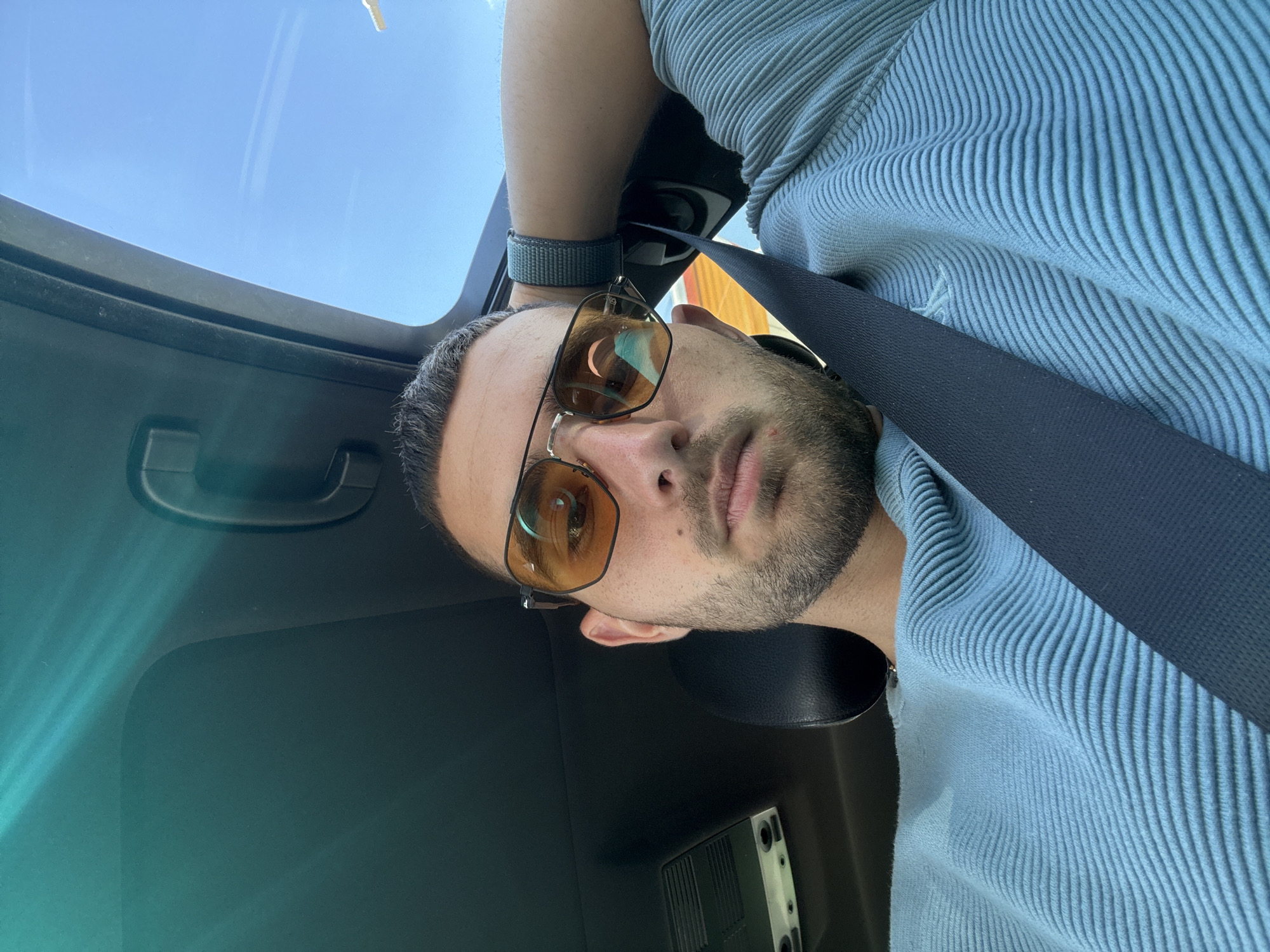 In Polsko Padarevo, Bulgaria, a man with short, dark hair and stubble is seated in a car, looking at the camera. He is wearing orange-tinted sunglasses and a light blue, ribbed long-sleeved shirt. A black seatbelt is fastened across his chest. His right arm is bent, with his hand resting on the back of his head. The car's sunroof is open, revealing a clear blue sky with faint clouds. The interior of the car, including the headliner and a grab handle, is visible.