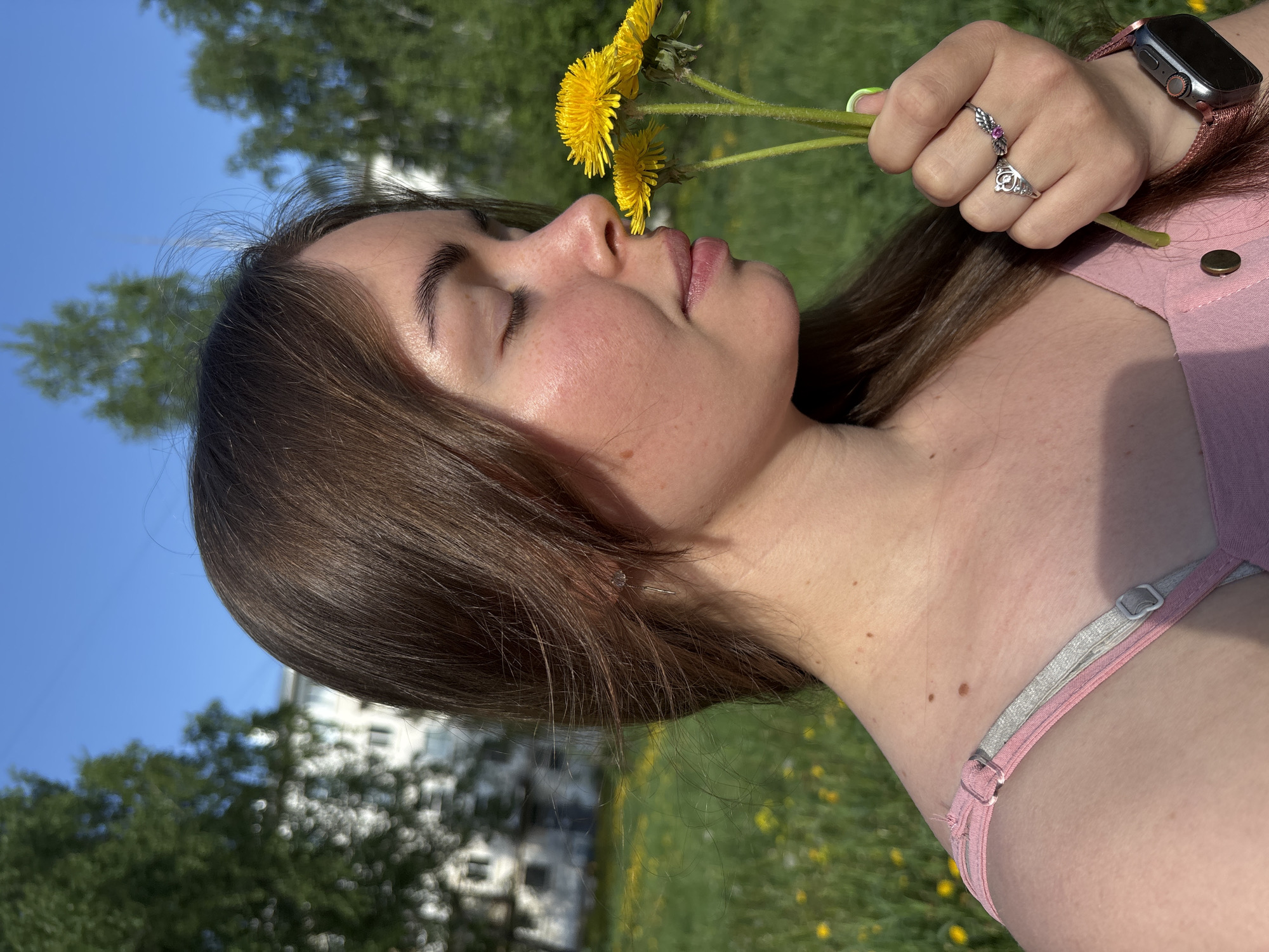 A young woman is holding dandelions to her nose with her eyes closed, standing in a grassy area, likely a park. She has brown hair and is wearing a pink top with thin straps. The woman is wearing a smartwatch and two rings on her right hand. In the background are trees, a building, and a clear blue sky. The environment appears to be a sunny outdoor location. There is no specific information to confirm the location as Izhevsk, Russia.