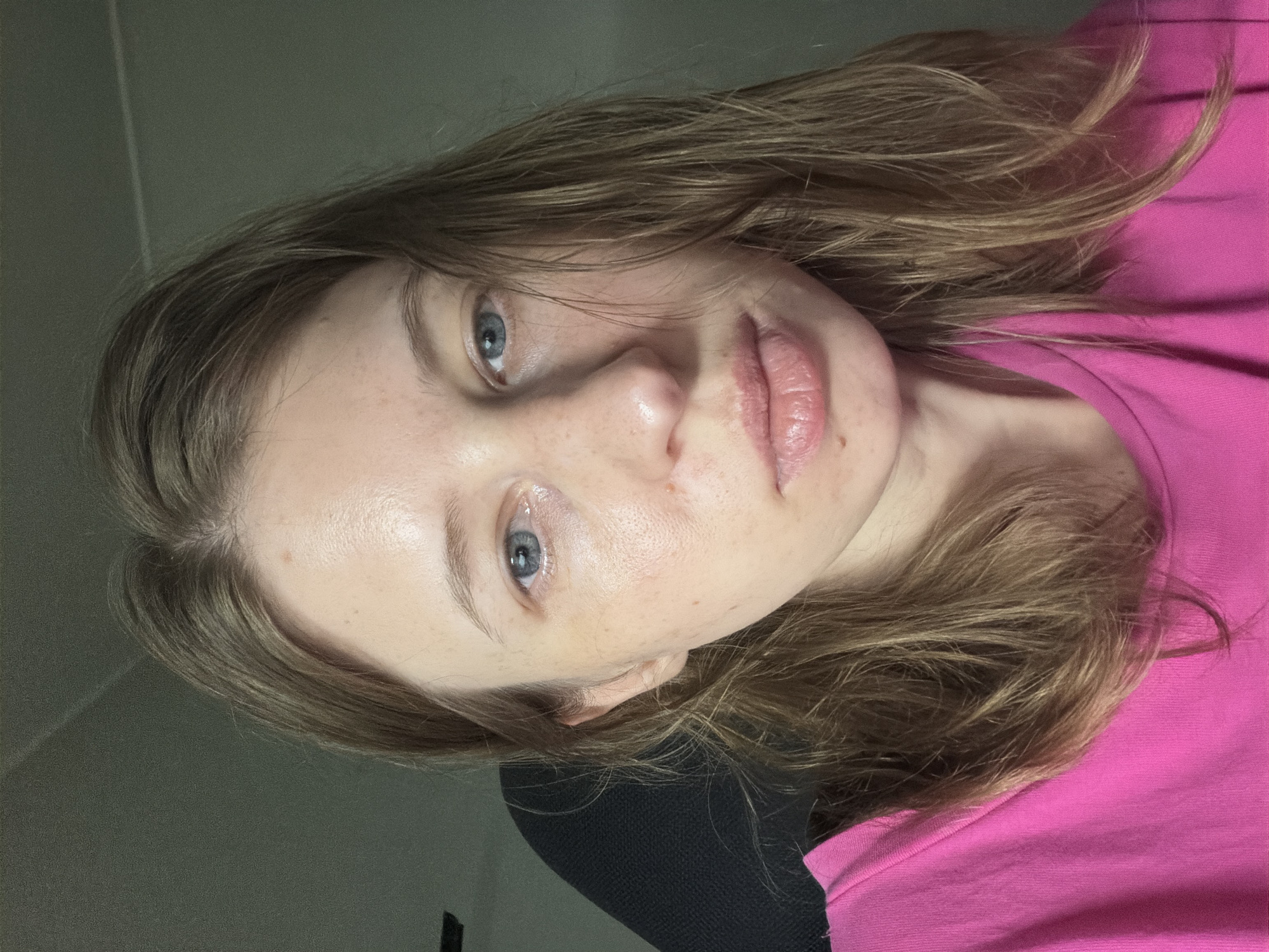 A young woman with shoulder-length brown hair and blue eyes is photographed from the chest up, wearing a bright pink t-shirt. She has fair skin with some freckles and appears to be in an indoor setting, possibly a home or office, given the plain background and a hint of a dark chair behind her. The lighting is soft, coming from the front and slightly to the side, illuminating her face and the texture of her skin. There is no discernible activity or interaction happening in the image, and the environment is nondescript.