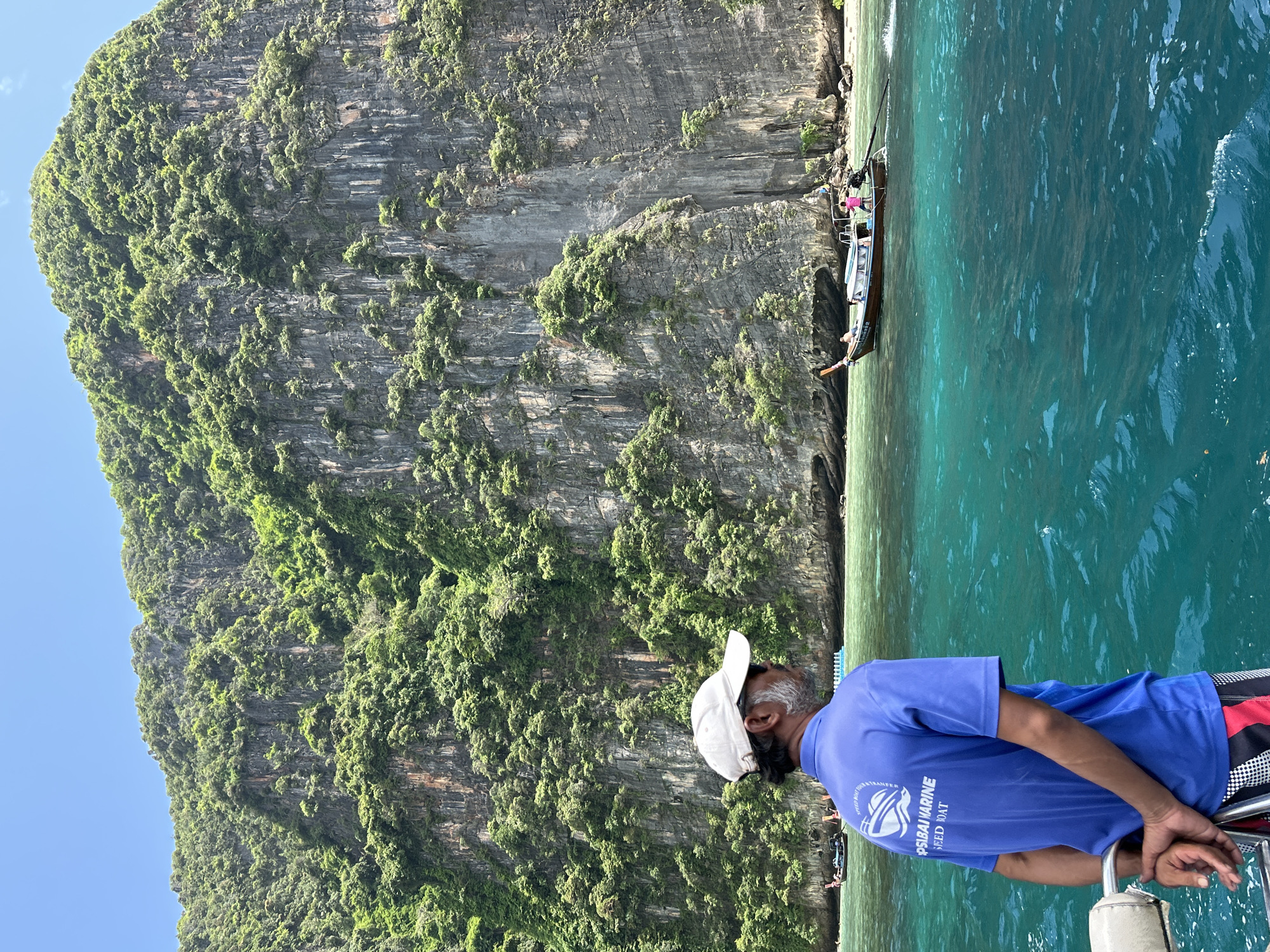 An adult male wearing a blue polo shirt with "PSIBAI MARINE SPEED BOAT" printed on the back and a white baseball cap stands on a boat, facing away from the camera. His hands are clasped behind his back. The boat is in the foreground, on a body of turquoise water. In the background, a sheer, tree-covered cliff face rises dramatically from the water. A longtail boat with several people on board is anchored near the base of the cliff. The sky is clear and blue. The scene is likely in Ao Nang, Thailand, known for its limestone karsts and island hopping tours.