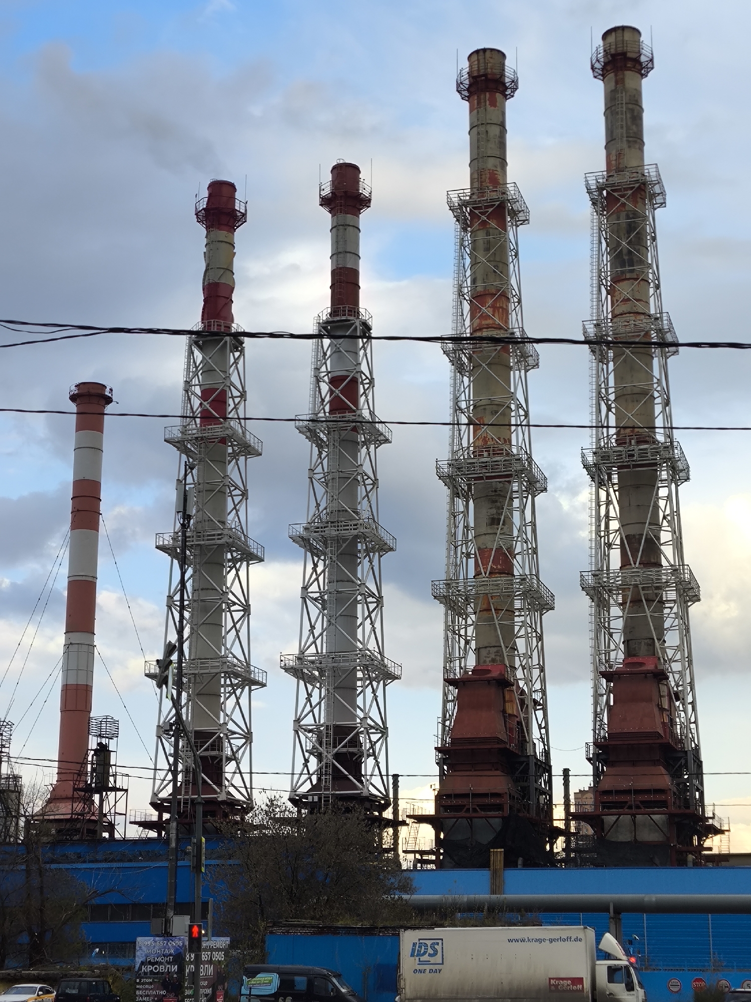 The image shows five tall, cylindrical industrial smokestacks, painted with alternating red and white bands at the top, supported by gray lattice-like steel frameworks. These are likely part of an industrial facility, indicated by the blue building structure beneath. Below, a street scene includes a white IDS truck with the website "www.krage-gerloff.de" printed on it, parked near a blue fence. A traffic light is visible on the left side of the image, showing a red signal. An advertising billboard displays text in Russian, and other vehicles are seen in the foreground. Overhead power lines cut across the top portion of the image, set against a sky with overcast clouds.