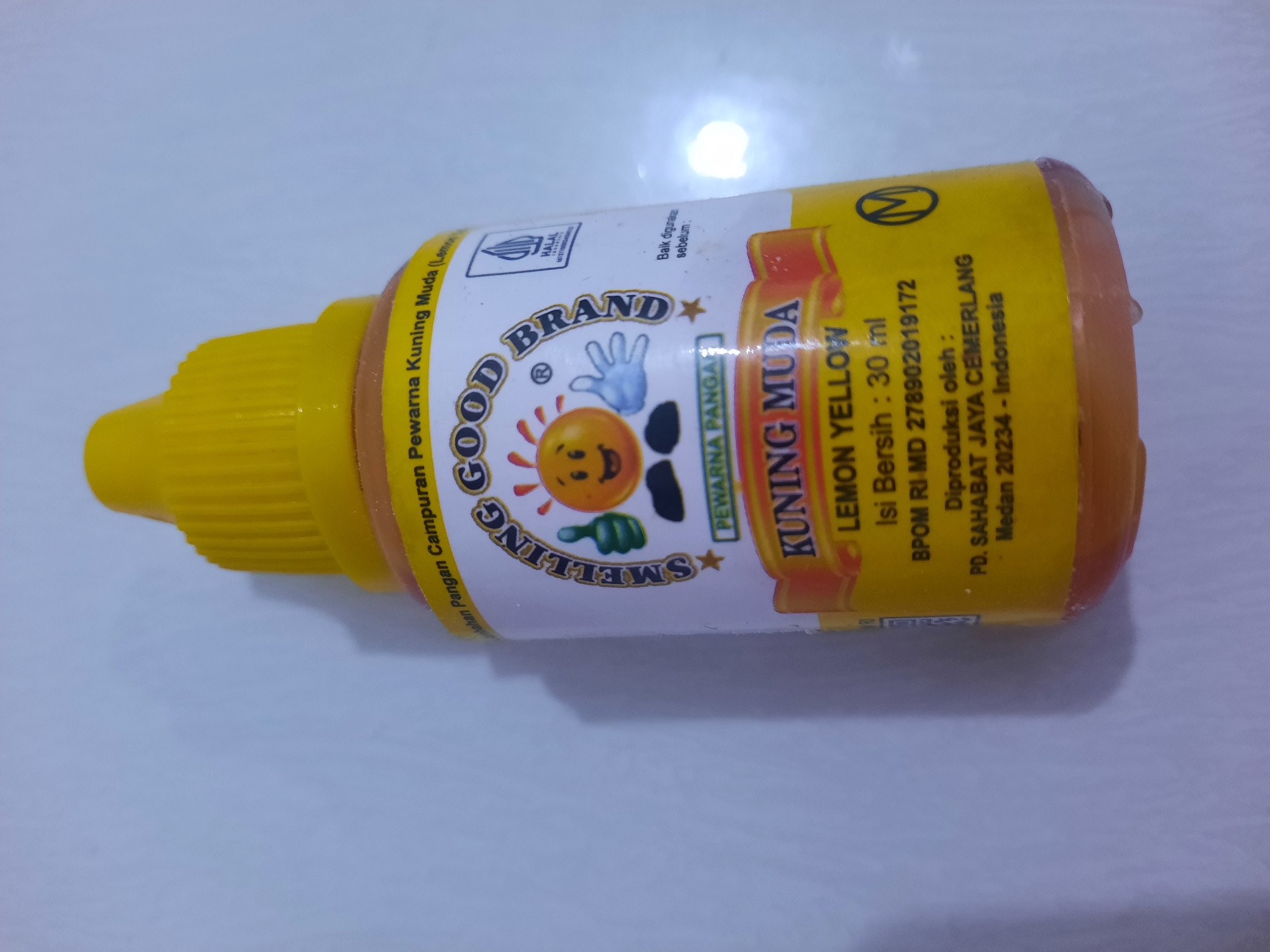 A yellow plastic bottle with a yellow cap is standing upright on a white surface. The bottle contains a liquid of a light orange-yellow color. The label on the bottle has various text and graphics. At the top of the label, there is text in Indonesian that translates to "Ingredients: Food Coloring Yellow Light (Lemon)". Below this, the brand name "SMELLING GOOD BRAND" is visible, with a cartoon face of a sun-like character and a hand giving a thumbs-up. There is also a "HALAL" logo. Further down, the label reads "KUNING MUDA LEMON YELLOW" and provides information about the net weight, BPOM registration number, and manufacturer details, including "PD. SAHABAT JAYA CEMERLANG Medan 20234 - Indonesia". The overall impression is a product shot of a food coloring bottle. There is no indication that this image was taken in Lhokseumawe, Indonesia.