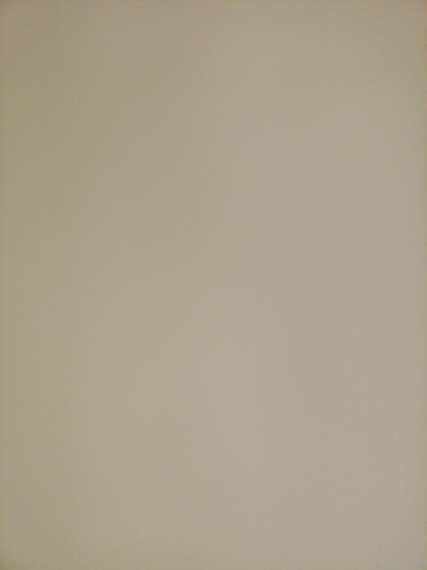 The image is a full-frame, dimly lit, neutral-toned photograph. The texture appears to be that of a flat, matte surface, possibly paper or a wall, with subtle variations in light and shadow that create a sense of depth without discernible features. There are no people, objects, or discernible environmental details present. The image is a uniform, light brown or beige color.