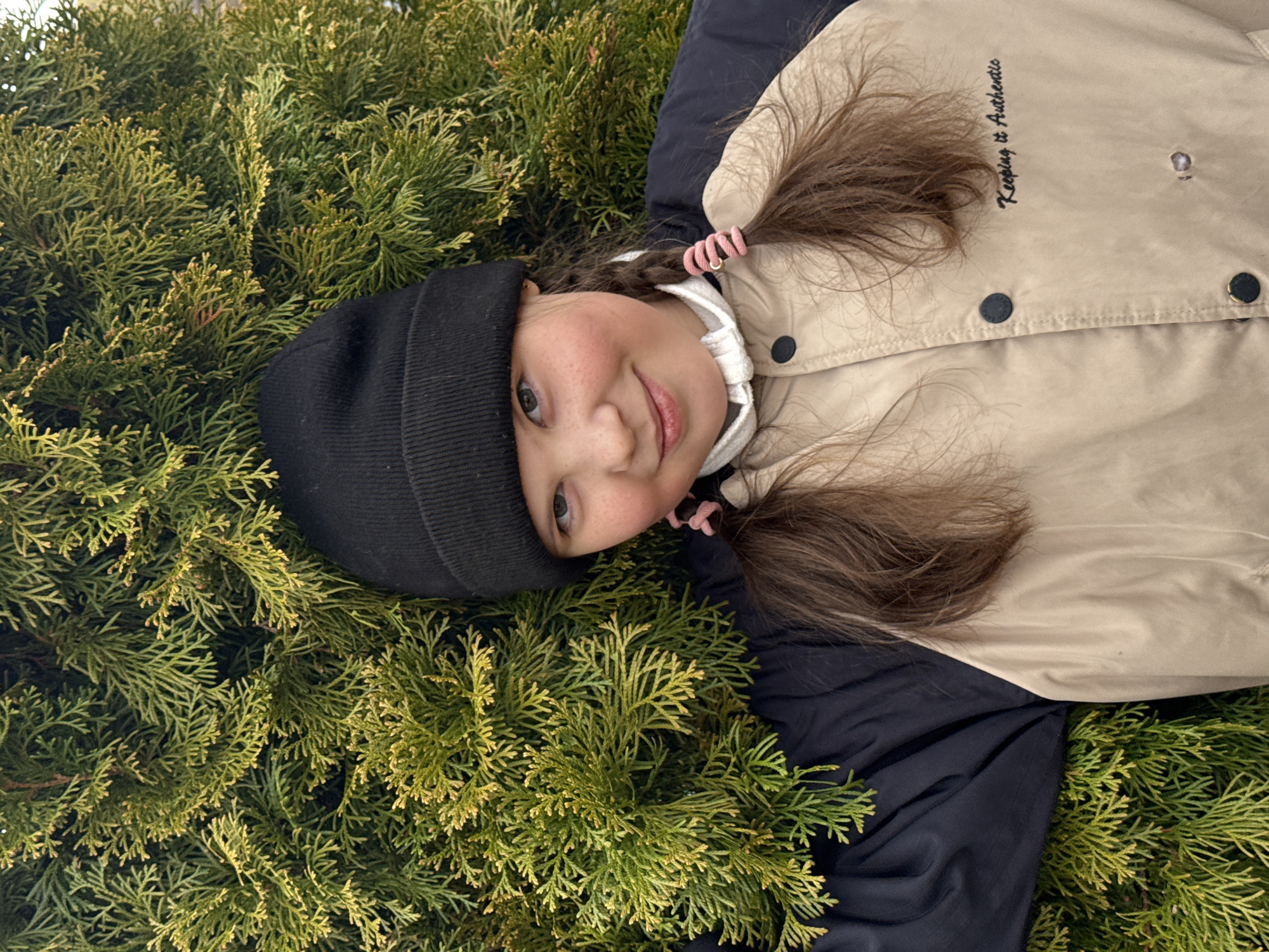 A young girl, appearing to be of primary school age, is the central subject of the image, lying down on her back amidst a dense bed of green coniferous foliage, likely thuja or cypress branches. She wears a black knit beanie and a two-tone winter jacket, which is beige on the upper half and black on the lower half and sleeves. Her brown hair is styled into two pigtails, each secured with a pink spiral hair tie.
The girl is looking directly at the camera with a gentle, slightly playful smile, her eyes sparkling. Her cheeks show natural freckles and a rosy blush. The lighting suggests it is daytime, providing an even illumination that highlights the vibrant green of the foliage and the details of her face and clothing. The attire indicates cool, possibly mild winter or early spring weather.
On the beige part of her jacket, above her right shoulder (viewer's left), the phrase "Keeping it Authentic" is embroidered in black cursive text. The overall scene conveys a feeling of calm and contentment. The location context for this image is Дойная, Russia.