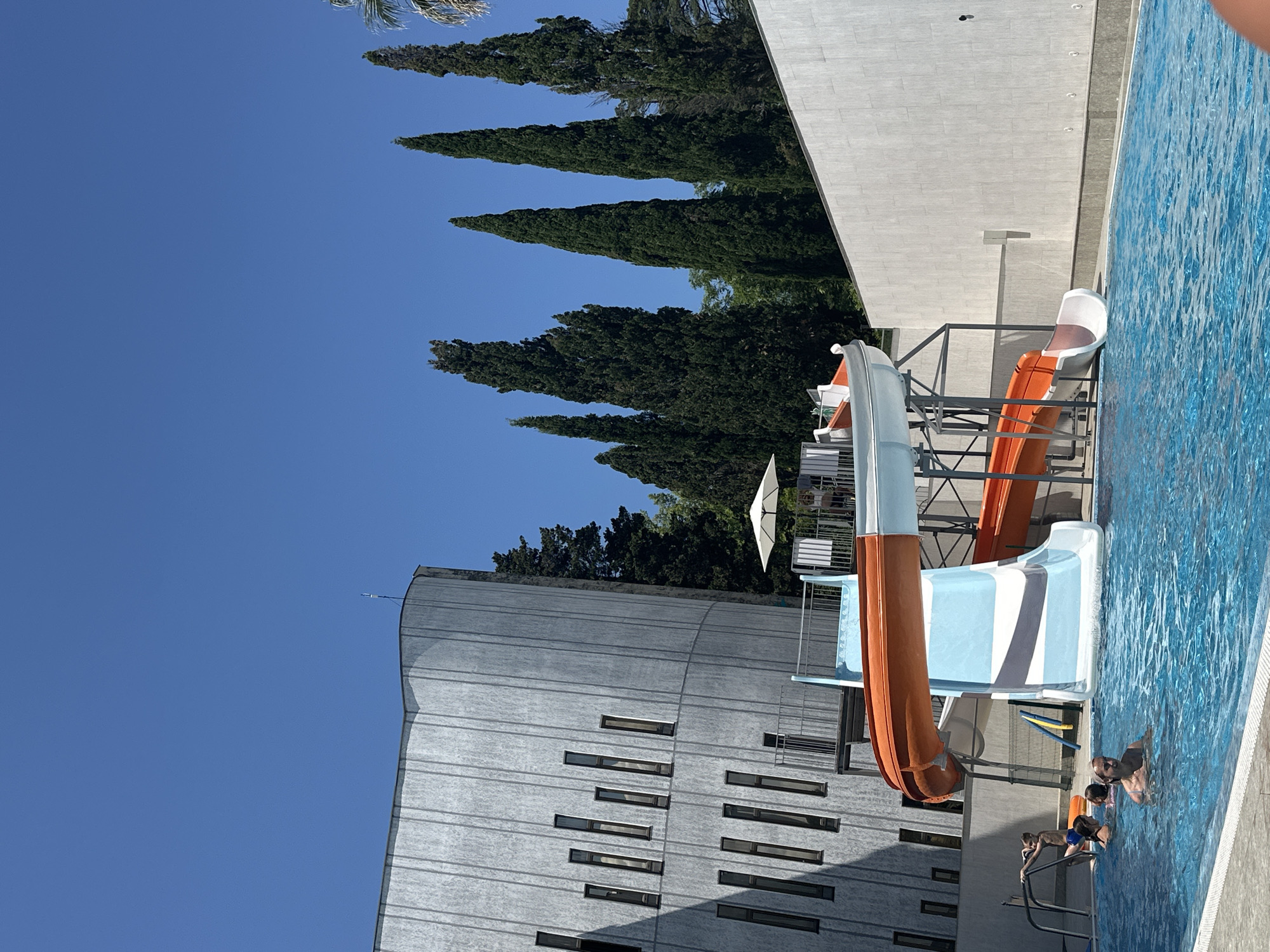 In Sochi, Russia, on a clear, sunny day, a blue swimming pool is occupied by several people. A water slide structure with orange and white slides is positioned next to the pool. To the left of the pool is a tall, modern building with a curved, textured gray facade and numerous narrow, rectangular windows. In the background, a line of tall, dark green cypress trees stands against the bright blue sky, with a white wall bordering the right side of the scene.