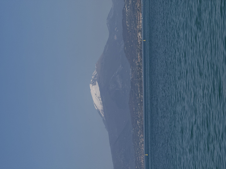 The image displays a wide view across Lake Garda, captured from the vicinity of Peschiera del Garda, Italy. The lower portion of the frame is filled by the lake's blue-green water, exhibiting minor surface ripples. Two yellow, cylindrical marker buoys are visible in the mid-distance on the water, positioned left-center and right-center. Beyond the lake, a densely populated strip of land with numerous structures forms a narrow band along the horizon. The background is dominated by a substantial mountain range; the most prominent and central peak is distinctly capped with snow. The sky above is clear and light blue, with atmospheric haze subtly muting the coloration of the distant mountains. No individuals, vessels, or specific actions are present in the observable scene.