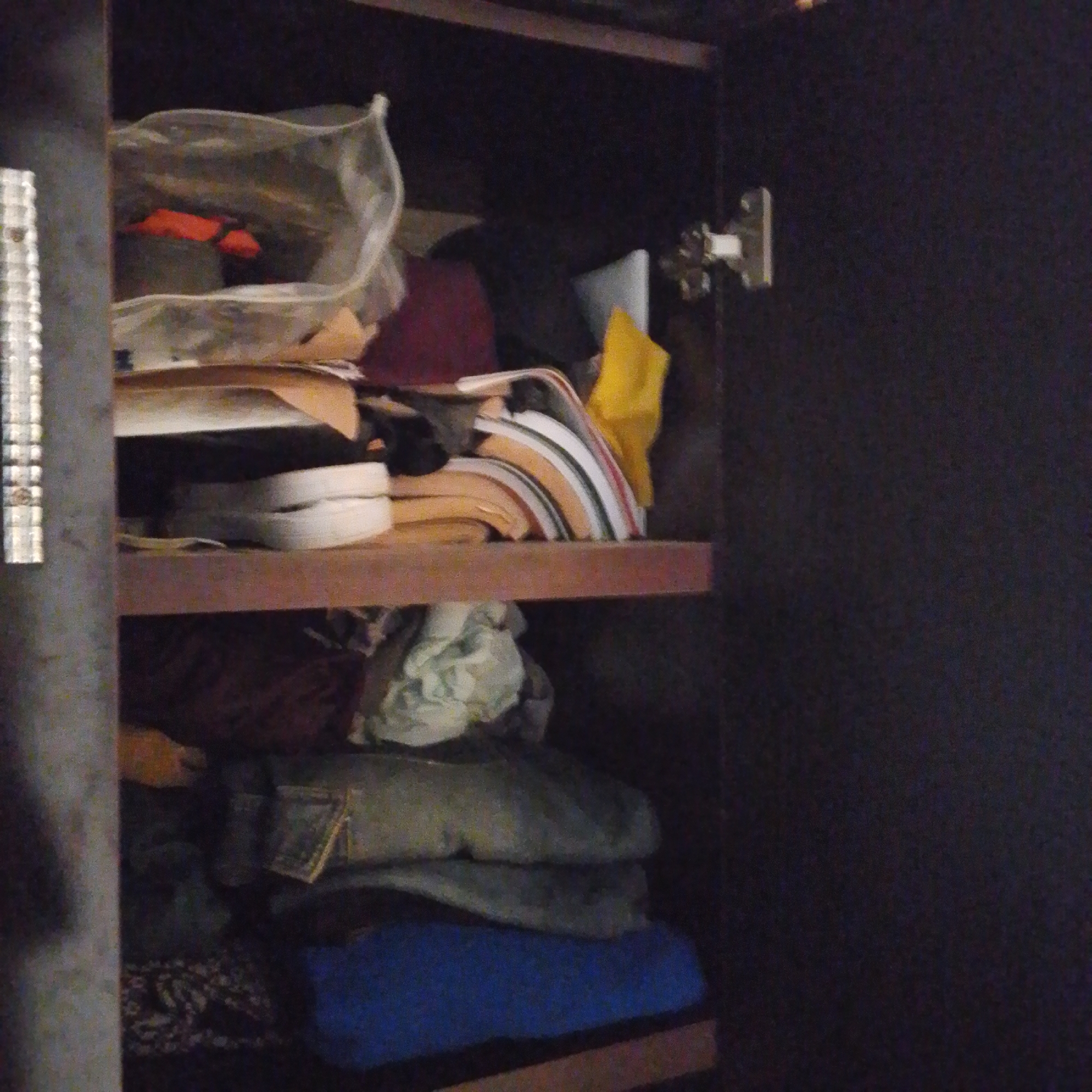 An interior shot of a dark brown wooden cabinet reveals two shelves stocked with various items. The top shelf holds a clear plastic bag containing red objects, possibly clothing, alongside a pile of folded fabric items with stripes of white, orange, and black, and a yellow piece of fabric. Below these, white shoes or sneakers are stacked. The bottom shelf contains more folded clothing, appearing to be a mix of darker fabrics and a crumpled light-colored textile. A bright blue fabric item is visible at the bottom of the cabinet, partly obscuring a patterned textile underneath. A metal hinge is attached to the right side of the cabinet door. The overall lighting is dim, with some light glinting off the metal hinge and the items on the shelves. The scene is within a residential setting in Jalingo, Nigeria.