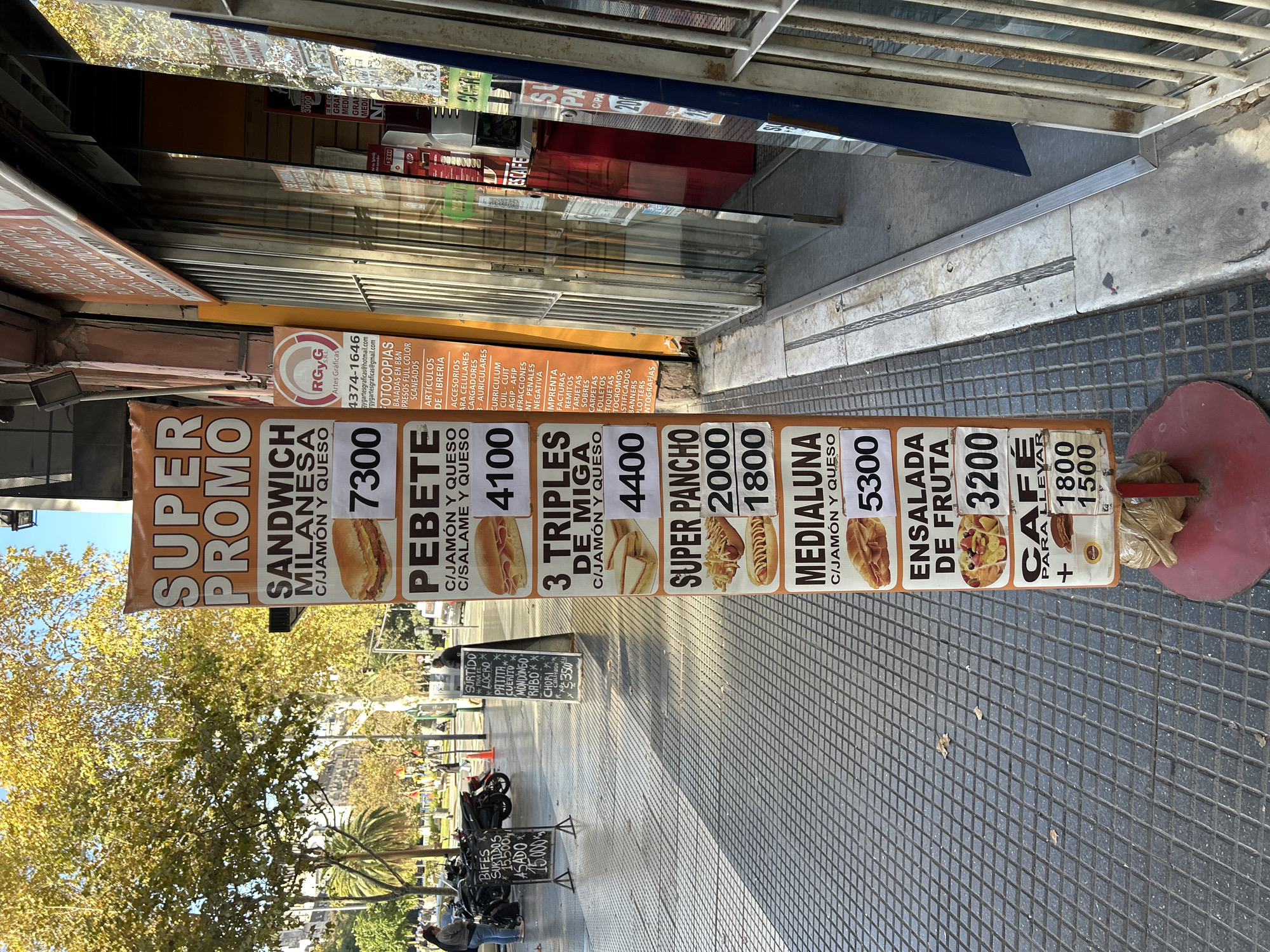The image shows a street scene in Buenos Aires, Argentina, with a tall, narrow advertising sign standing on the sidewalk in front of a commercial storefront. The sign displays a menu of food items with prices, including sandwiches, salads, and coffee. A smaller chalkboard next to parked motorcycles offers other food items and prices. The storefront is protected by a metal roll-down security gate. A tree with yellow leaves and other street furniture are visible in the background, along with several pedestrians. A large orange sign is seen in the background.