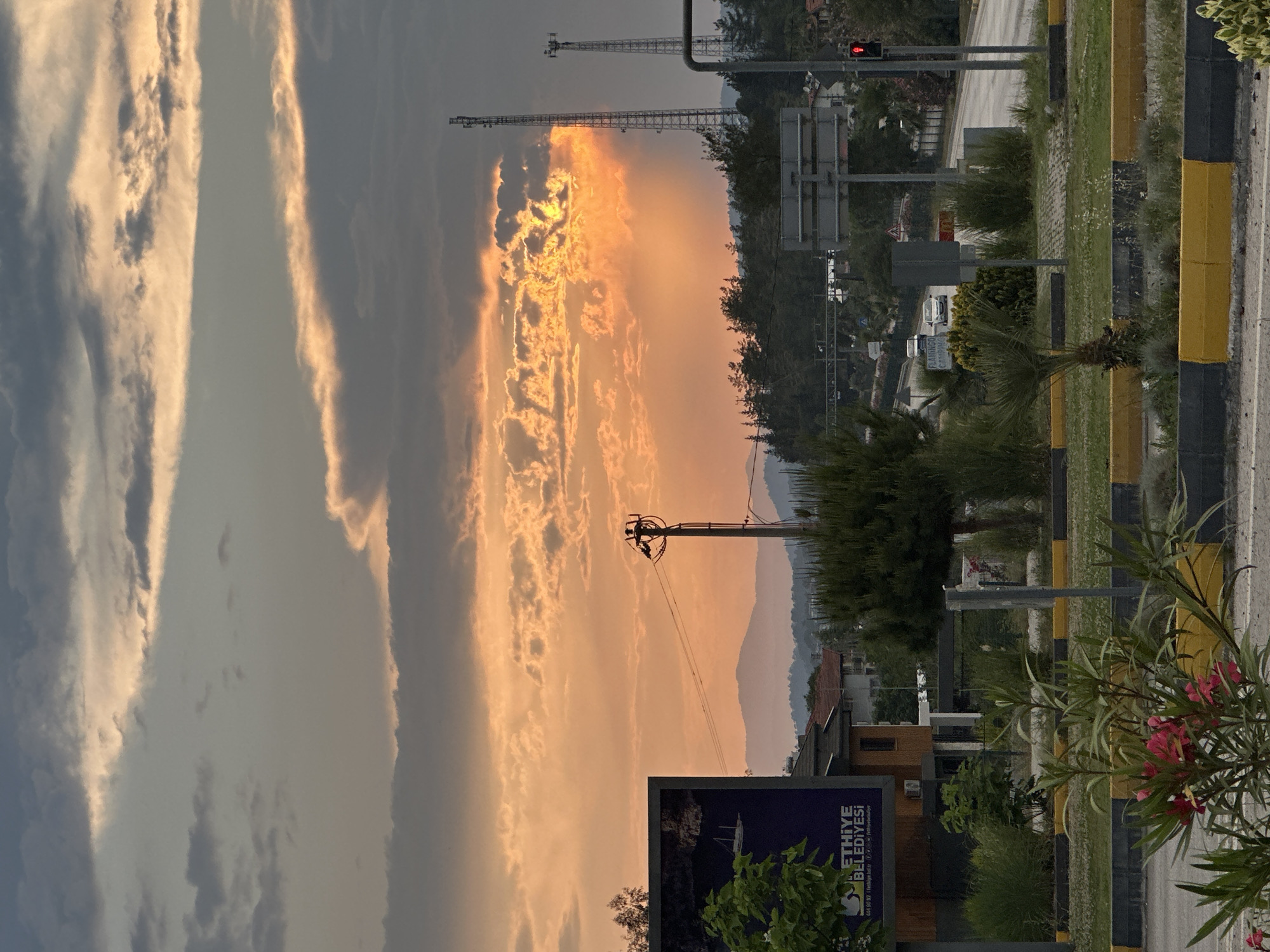 The image shows a cityscape in Fethiye, Turkey, during a sunset or sunrise, with a vibrant sky dominated by shades of orange and grey clouds. Two communication towers stand silhouetted against the bright sky. An electrical pole with attached wires is visible, connecting to nearby buildings. In the foreground, there's a road with a dividing strip marked with yellow and black poles, and a variety of greenery, including trees, bushes, and oleander plants with pink flowers. A large advertisement board featuring the words "FETHIYE BELEDIYESI" (Fethiye Municipality) in Turkish is present. In the distance, mountain ridges create a backdrop to the urban scene. A traffic light appears to be red.