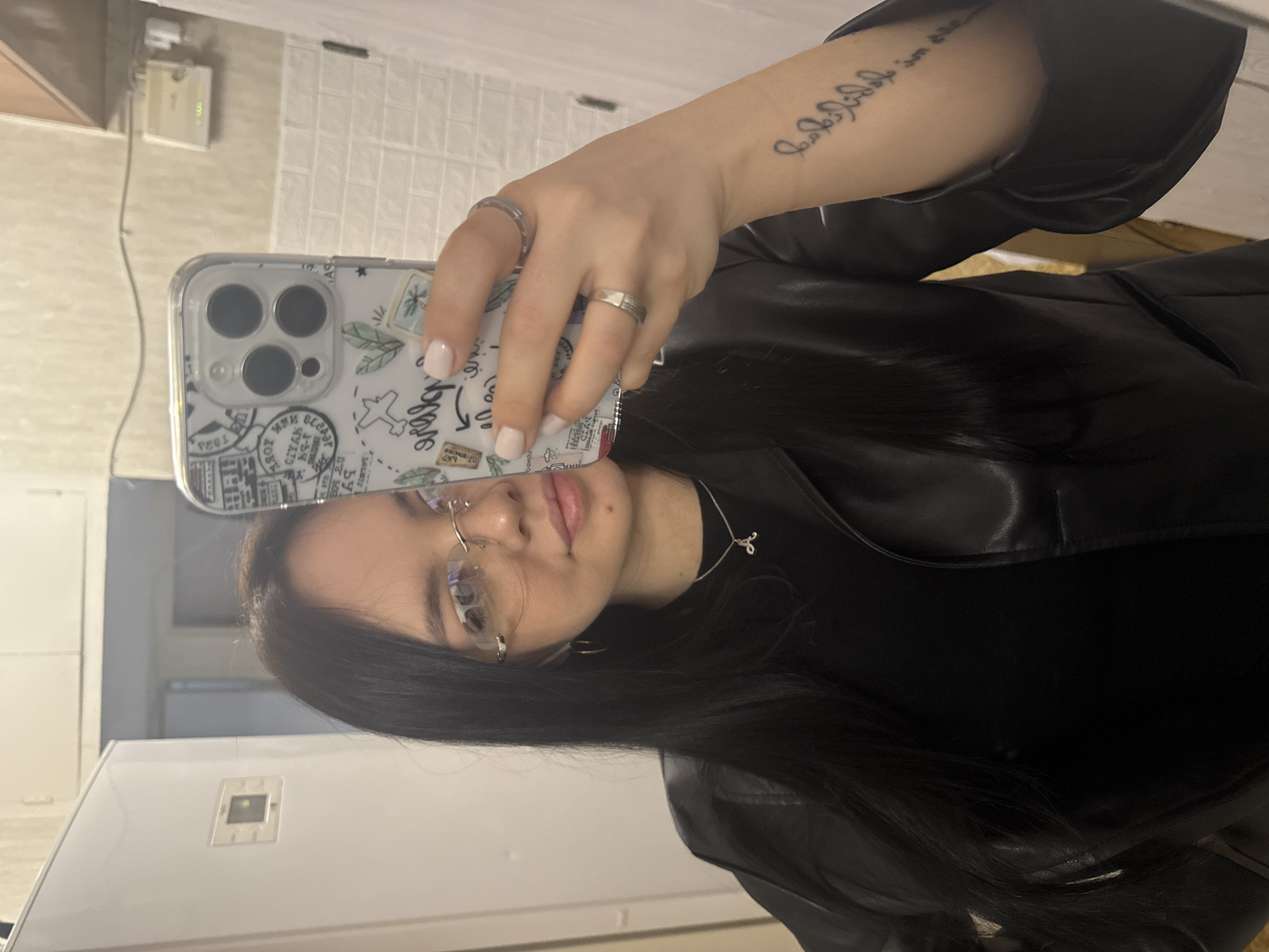 The image depicts a woman taking a selfie in what appears to be an apartment. The location is unspecified.
The woman is wearing a black shirt and a black leather jacket. She has long, dark hair and is wearing glasses. She is holding a phone in her left hand to take the photo. The phone has a patterned case. The woman is wearing rings on the fingers of her left hand. She has a tattoo on her left forearm.
The background includes a kitchen area with white cabinets, a white refrigerator, and a white brick-patterned wall. A small electronic device is visible on the wall.