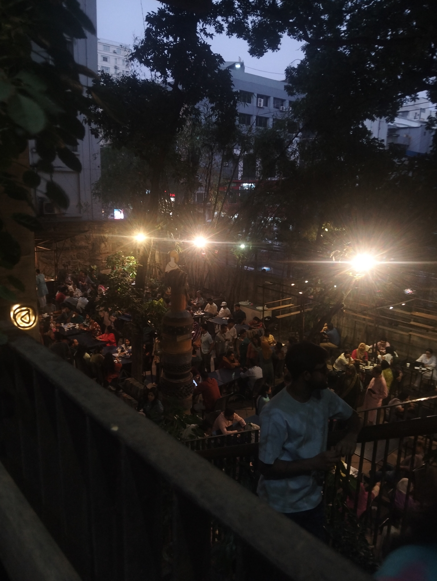 The image shows a crowded outdoor restaurant in Dhaka, Bangladesh, illuminated by bright overhead lights. Numerous people are seated at tables, appearing to dine. In the foreground, a man in a light-colored shirt leans against a railing, partially obscuring the view. The restaurant is surrounded by trees and buildings, suggesting an urban setting.