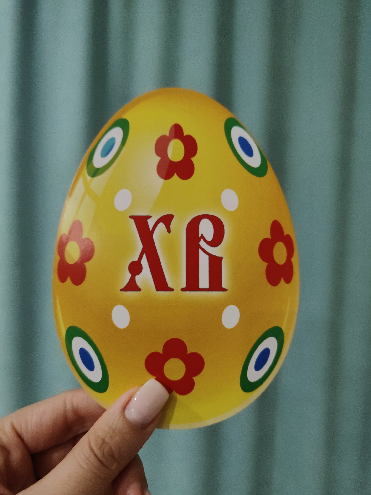 A hand holds a yellow Easter egg decorated with red flowers, blue and green circles, and the red Greek letters "ΧΡ" against a turquoise curtain. The scene is indoors. The location is unknown.