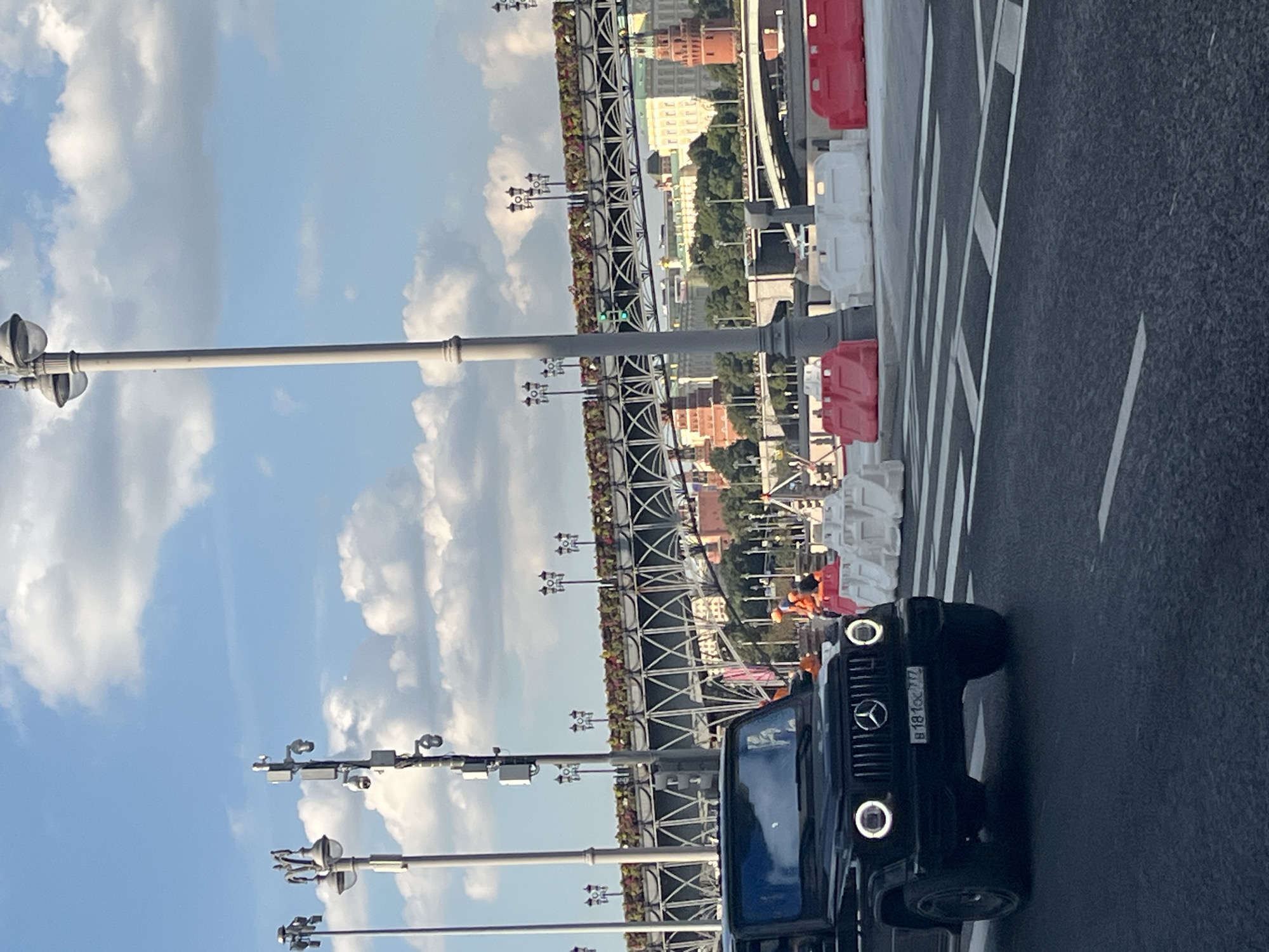 The image shows a black Mercedes-Benz G-Class SUV driving on a road in Moscow, Russia. In the background, there is a bridge decorated with flowers, and beyond that, the Kremlin can be seen with its distinctive red towers. The sky is blue with some white clouds. The road has white lines marking lanes and a pedestrian crossing. There are temporary barriers on the side of the road, some red and some white, and some construction workers in orange vests are visible near them. The overall scene suggests a sunny day in a busy urban environment.