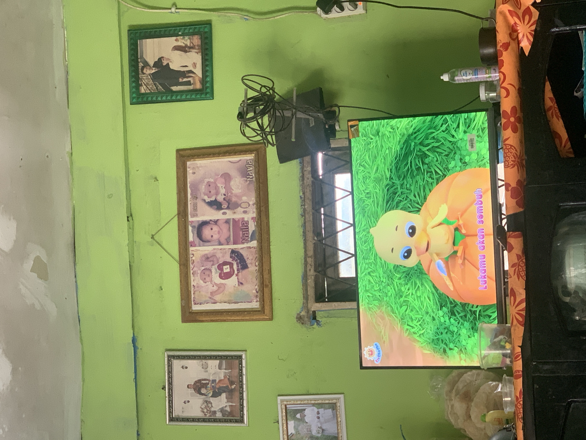 The image captures a domestic scene, likely a living room, within a residence in Palembang, Indonesia. The walls are painted a vibrant lime green, showing signs of wear and tear with chipped paint and exposed plaster. Several framed photographs hang on the wall. On the left, a framed photograph depicts a family, possibly a wedding portrait. Above it, another framed photo shows a baby. To the right, a green-framed photograph displays a couple, and next to it, a golden-framed picture features two children.
In the foreground, a television screen displays a cartoon of a yellow chick sitting on an orange surface, with green grass in the background. Below the screen, a dark-colored cabinet is visible, covered with an orange and red patterned cloth. On the cloth, several items are placed, including a bottle of "My Baby" brand antiseptic liquid, a small jar, and a dark mug. To the left of the television, a stack of flat, round crackers and a plastic container are also present. Wires are seen connected to a device mounted on the wall behind the television, which appears to be an antenna or similar electronic equipment. A white electrical outlet with a switch is mounted on the wall to the right. The overall impression is of a lived-in space, possibly indicating a modest dwelling.