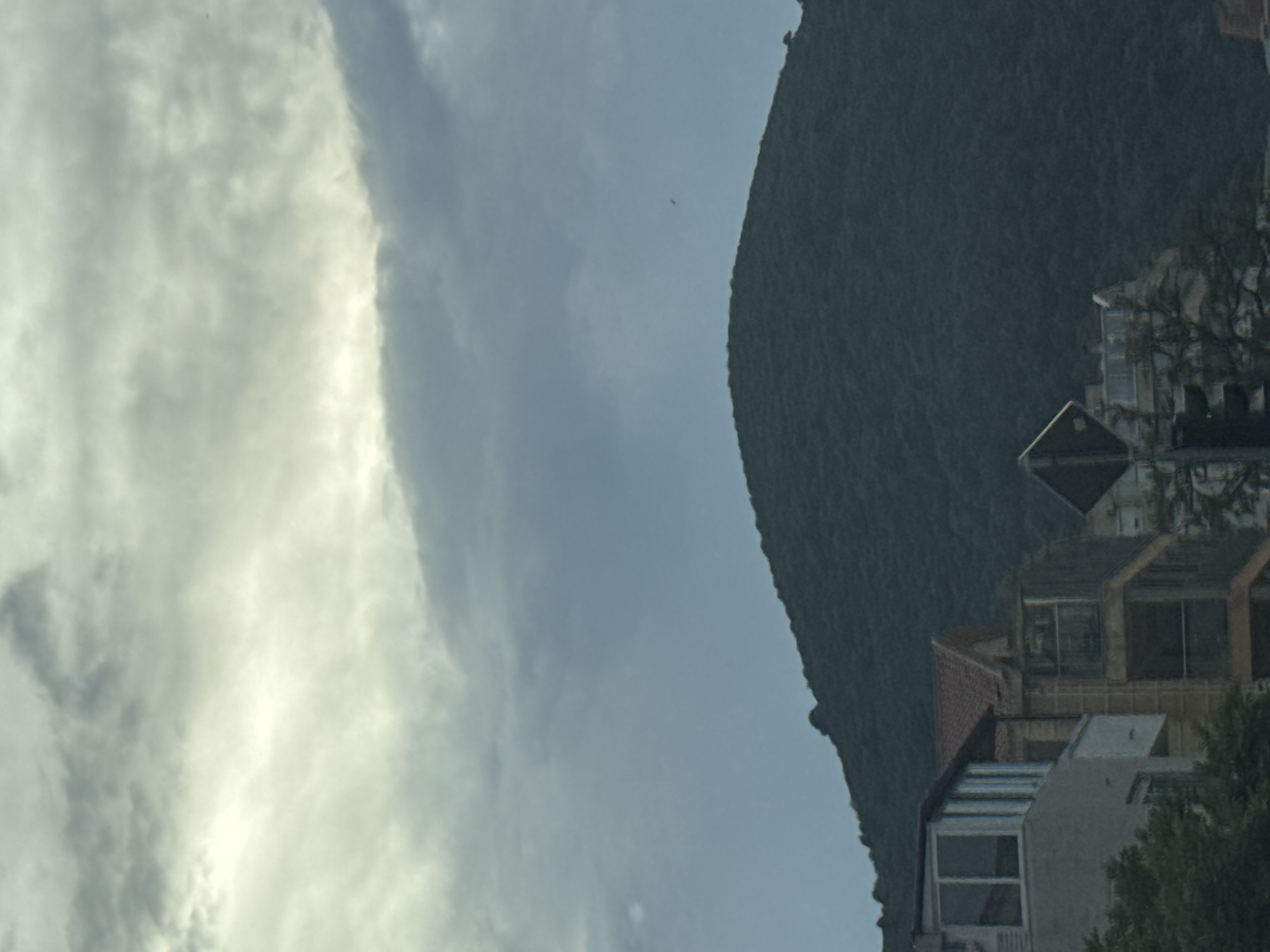 The image captures a view from a street in Budva, Montenegro, looking towards a forested hill. The sky is overcast with layers of gray and white clouds, with a brighter band of light breaking through from the upper left. In the foreground, several multi-story buildings with balconies and windows are visible. The architecture suggests a residential or commercial area. A street sign with a diamond shape is present on the left side, indicating a road junction or warning. The hillside is covered in dense green vegetation, appearing as a dark, textured mass against the sky. The overall impression is a somewhat muted, cloudy day in a coastal town.