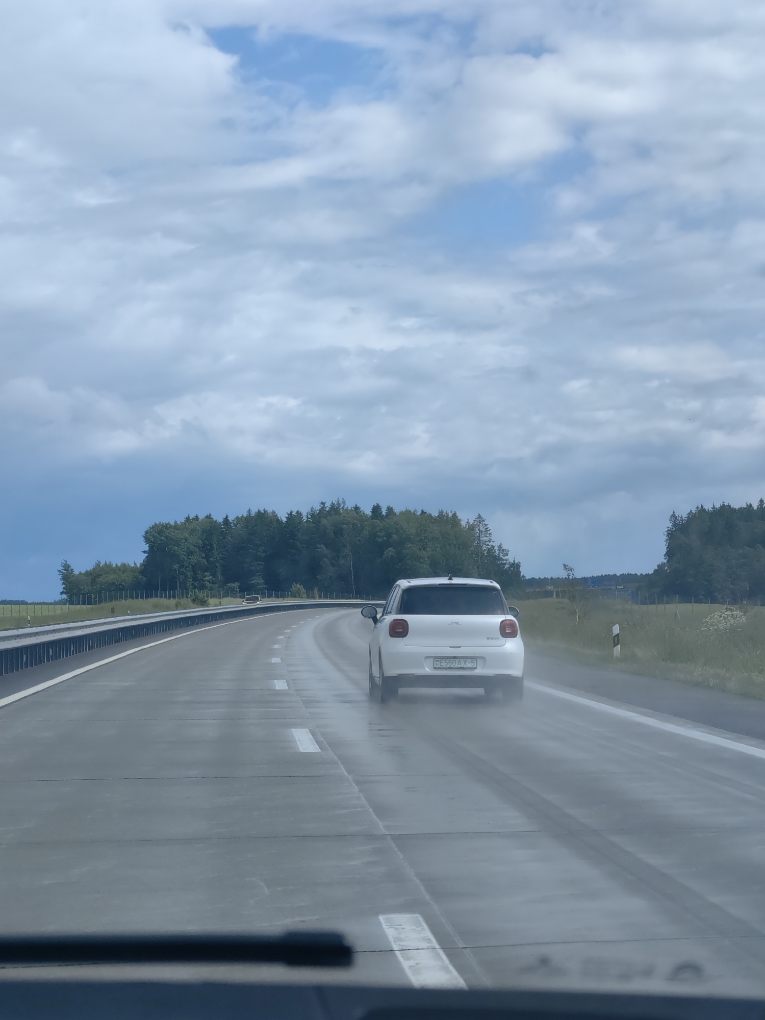 The image depicts a wet highway in the vicinity of Путчынскі сельскі Савет, Belarus, under a cloudy sky. A white car is driving on the highway, leaving a spray of water in its wake. The road is wet and reflective, with white lane markings visible. Trees line the right side of the highway, and a metallic guardrail is present on the left. The perspective suggests the photo was taken from another vehicle.