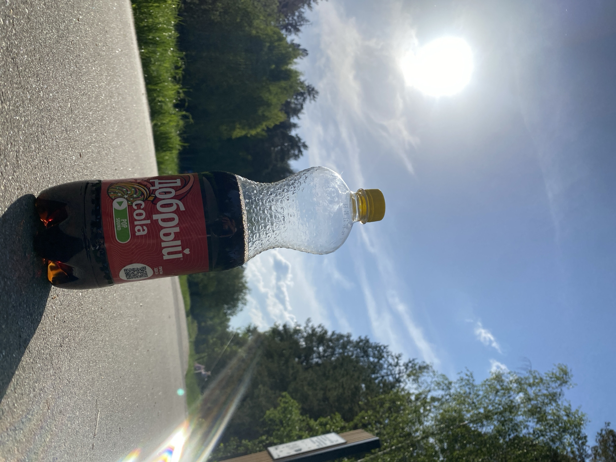A bottle of Dobry Cola lies on a paved surface, likely a sidewalk or road, on a sunny day. The sky is blue with scattered clouds, and the sun is bright and high in the sky. Trees line the background. The location is Unknown, Unknown.