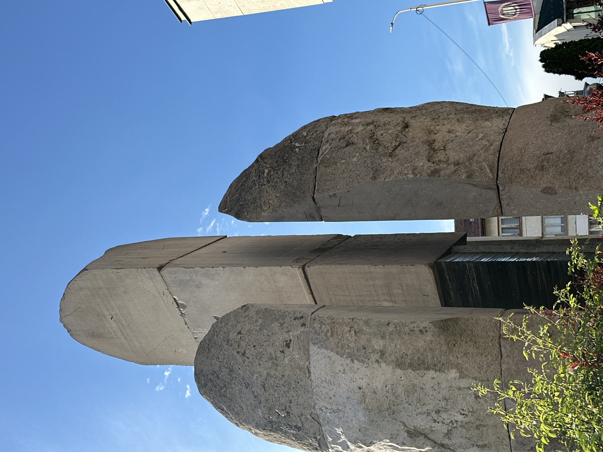 The image shows the Monument to Vasil Levski in Lovech, Bulgaria, from a low angle. The monument is constructed from large, grey stone blocks. Sections of the monument are separated by thin gaps. In the background, buildings and a flag are visible. Some greenery is present on the right. The sky is clear and blue.