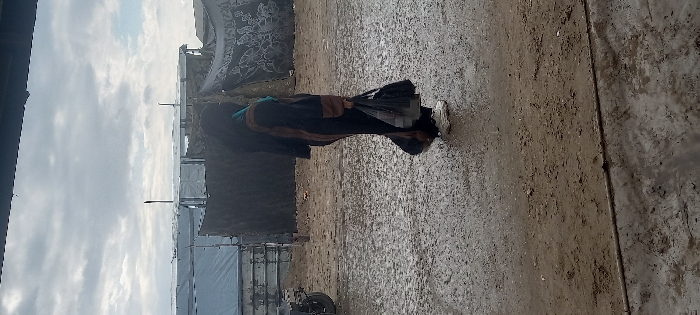 The image depicts an outdoor scene in Khan Yunis, Palestinian Territory, under an overcast sky. The ground is predominantly wet, muddy earth with numerous small puddles and granular debris, suggesting recent precipitation.
A lone female figure is positioned in the mid-ground, captured in profile, walking from left to right. She wears a dark, full-length abaya with brown trim on the sleeves, a dark head covering, and white athletic shoes. A teal-blue garment or scarf is visible around her neck area beneath the abaya. In her left hand, she carries two dark-colored, soft bags.
To the left of the woman, partially obscured, is the rear section of a dark-colored, two-wheeled vehicle, possibly a motorcycle or bicycle. The background features a dense collection of makeshift shelters or tents, constructed from various materials including silver tarpaulins, corrugated sheets, and dark fabric. One shelter on the right exhibits a patterned dark fabric wall. Multiple vertical poles are visible, supporting these structures. The upper foreground is shadowed by a dark, indeterminate overhead structure, likely an awning or roof edge.