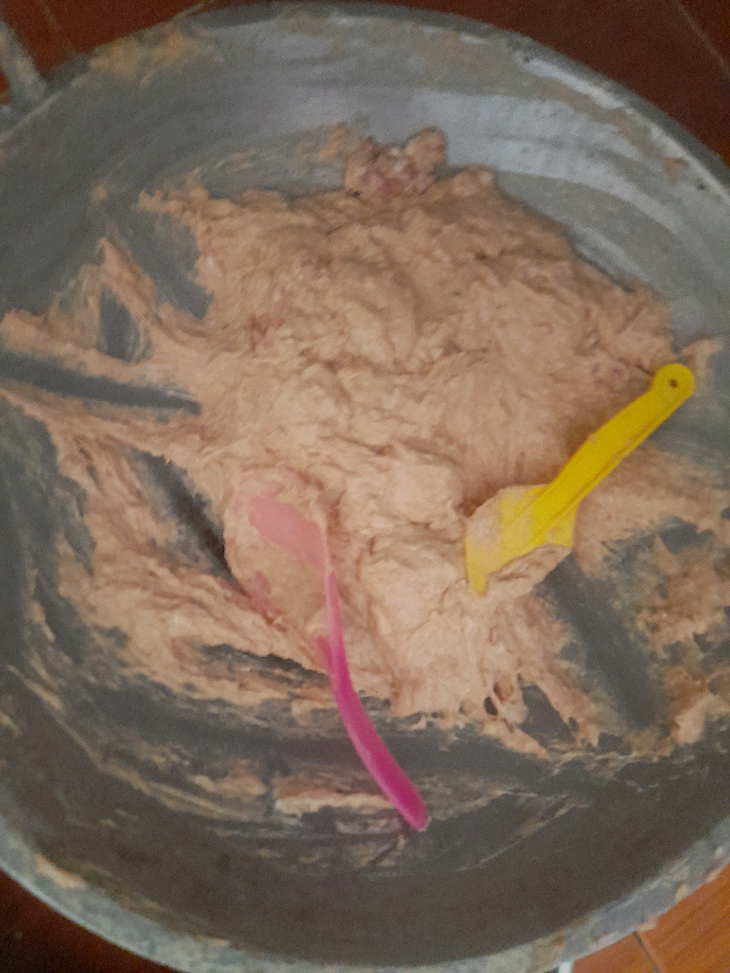 A close-up, overhead shot shows a large, dark gray metal bowl filled with a light brown, dough-like substance. Two plastic utensils are partially submerged in the mixture: a bright yellow spatula on the right and a pink spatula on the left. The bowl appears to be on a reddish-brown surface, possibly a wooden table. The background is blurred and out of focus, but the overall scene suggests a food preparation activity.