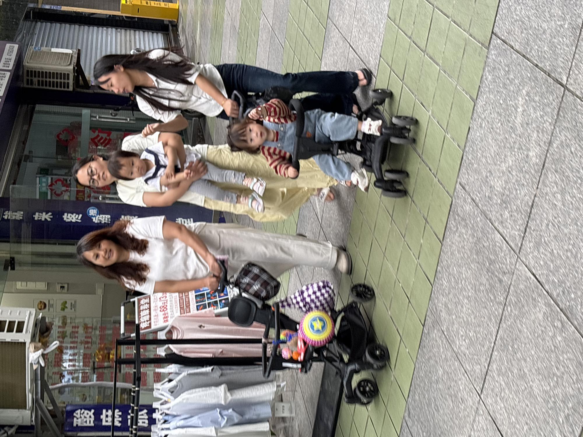 The image depicts a street scene with four people and two strollers. In the foreground, a woman with long, brown hair is pushing a black stroller adorned with a purple and white checkered fabric and a yellow and blue toy. Behind her, a second woman, wearing glasses and a light-colored top, is holding a baby. To her left, a third woman, with long dark hair, is leaning over and looking at a child in a stroller. The child, wearing a striped shirt and blue overalls, is looking towards the camera. The scene appears to be outdoors, possibly a sidewalk, with a tiled ground composed of green and gray squares. In the background, there are shop fronts with visible signage. One sign, partially visible, reads "疼痛康养堂" (Pain relief and wellness hall) and other Chinese characters. The lighting suggests it might be daytime. The people's expressions are neutral. The overall impression is a family outing in a commercial or public area.
