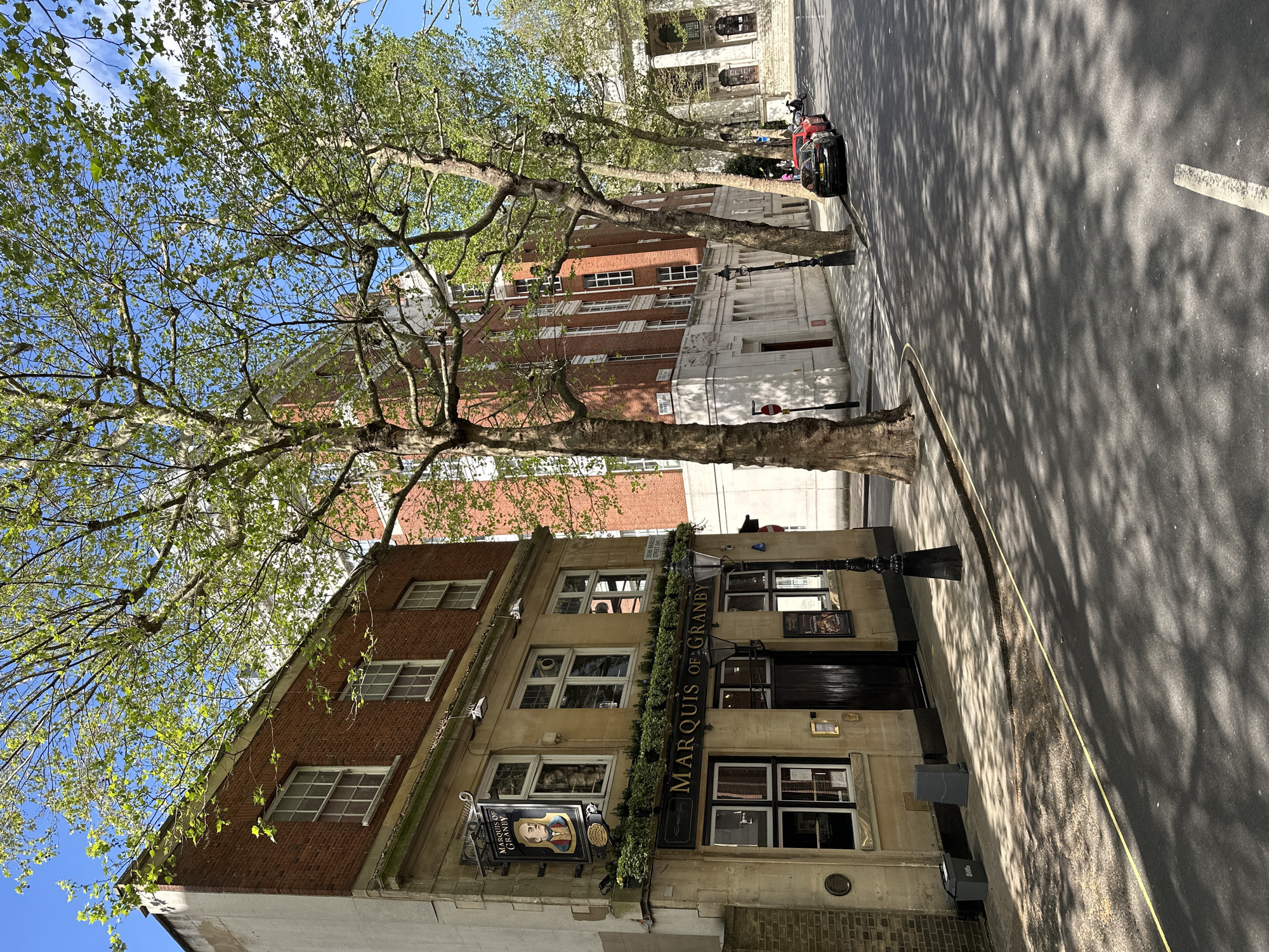 The image shows the Marquis of Granby pub in London, United Kingdom. The pub is a multi-story building with a brick facade on the upper floors and a stone facade on the ground floor. There are several windows on each floor, and a sign with the pub's name hangs on the front of the building, adorned with greenery. The image includes the tree that stands near the building.
Adjacent to the pub is a street with cars parked along the side. Shadows from the trees cast across the road surface. In the background, other buildings can be seen, including a larger structure with white columns.
The sky is blue, and the overall impression is that of a sunny day.