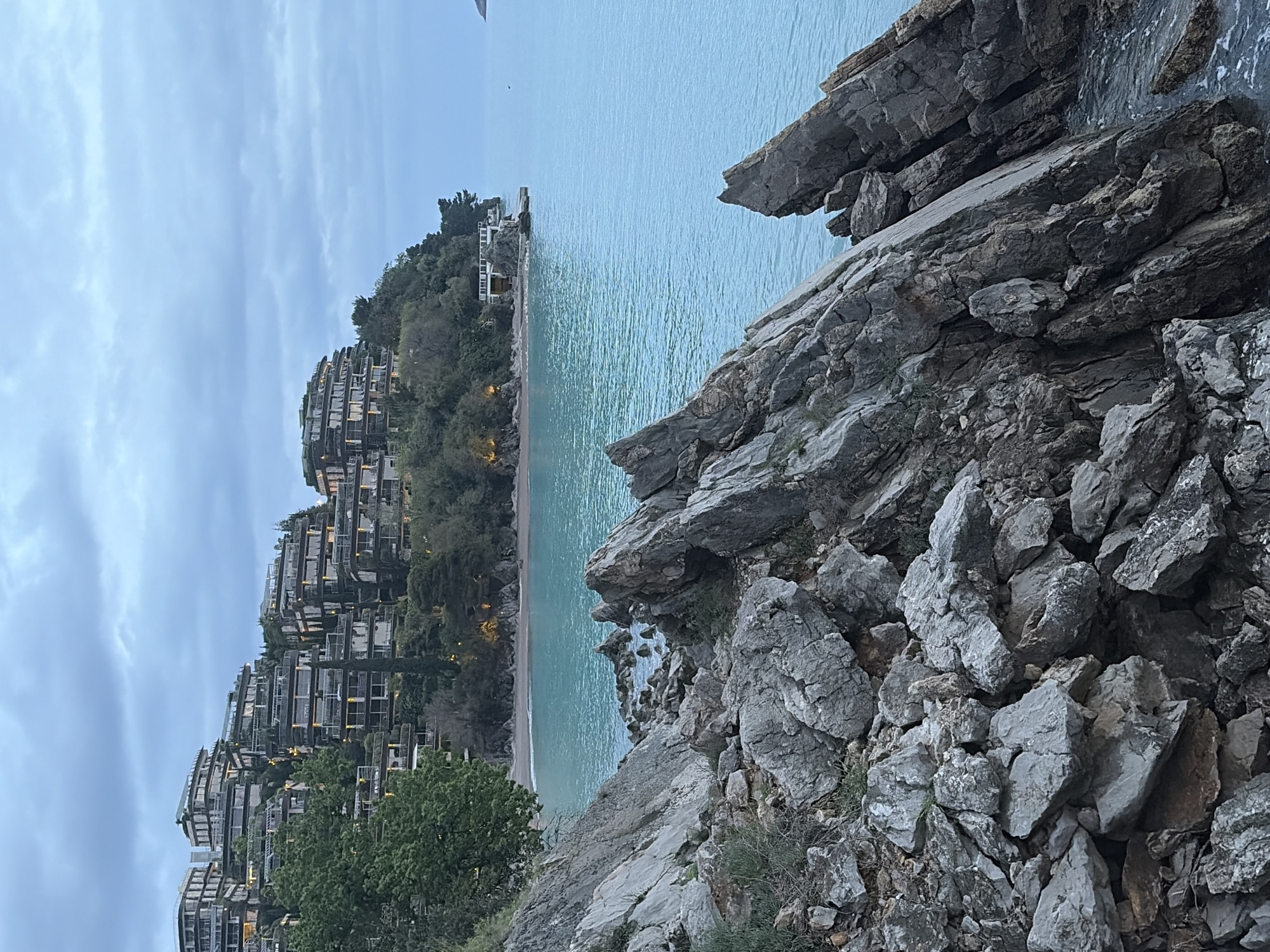 The image captures a coastal scene in Budva, Montenegro, showing a view of a rocky coastline and the Adriatic Sea. The foreground is dominated by large, gray rocks and cliffs, which appear rugged and uneven. Vegetation, including small shrubs and patches of green, are scattered among the rocks.
In the mid-ground, there is a stretch of beach along the water's edge. Beyond the beach, a series of terraced buildings or apartments are built into the hillside, suggesting a residential or resort area. The buildings are multi-story and have balconies, and the architecture suggests modern construction.
The sea is calm and a light turquoise color, extending towards the horizon under an overcast sky. The overall scene suggests a combination of natural coastal beauty and human development in a seaside resort setting.