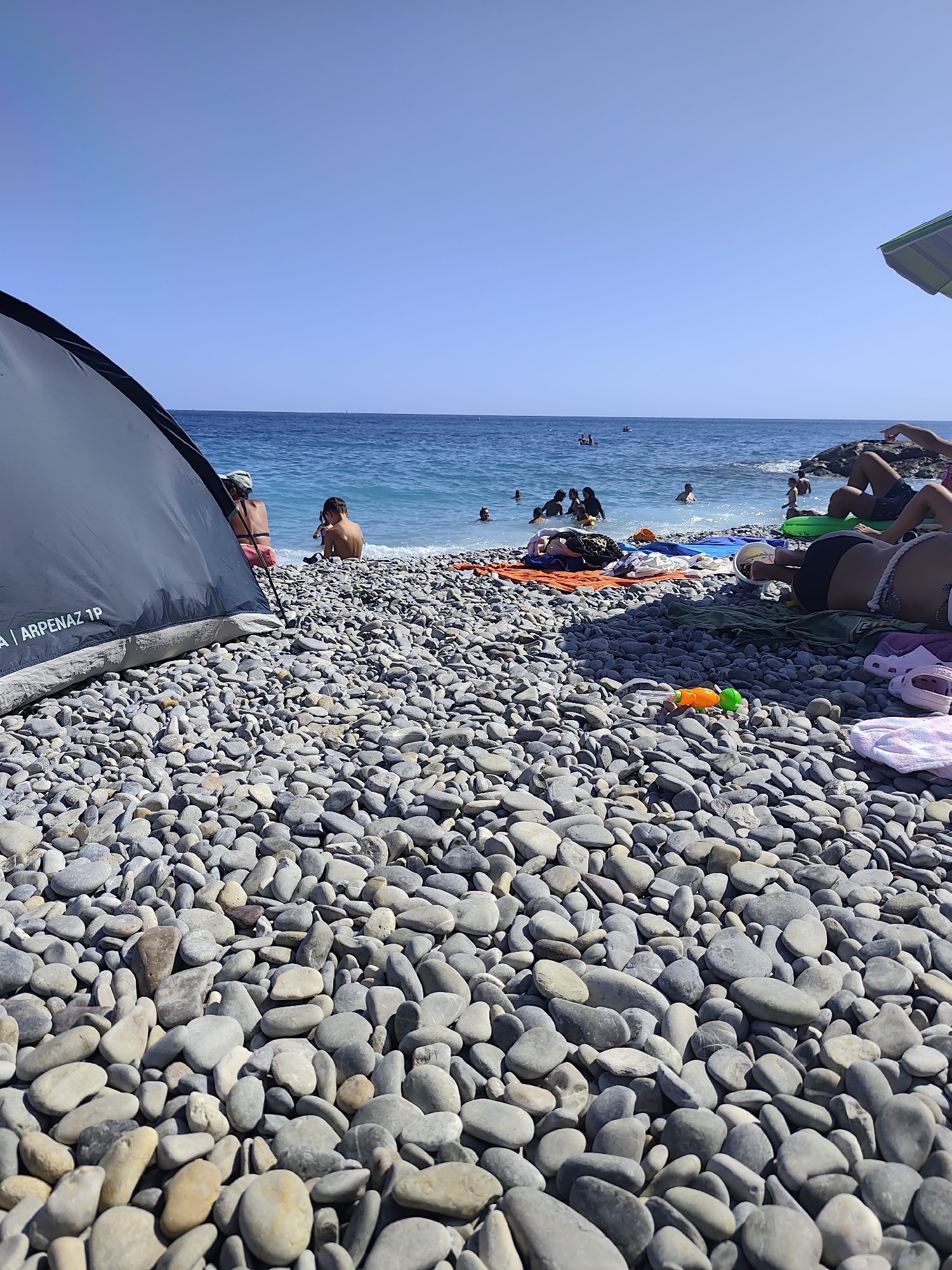 The image depicts a crowded pebble beach on a sunny day. In the foreground, a grey tent is set up on the left, and a collection of beach towels and a green inflatable ring are spread out on the pebbles. A person's legs and torso, wearing black shorts and a rope detail, are visible on the right, lounging on the beach. A small orange and green water gun lies on the pebbles in front of them.
In the middle ground, several people are in the water, swimming and enjoying the sea. The water is a clear blue, with waves gently breaking on the shore. Beachgoers are visible sitting and standing on the water's edge.
The background shows a clear blue sky and the vast expanse of the sea. A faint sailboat is visible on the horizon. The rocky coastline is visible in the distance. The overall scene suggests a popular summer day at the beach in Nice, France, characterized by vibrant blue waters and a lively atmosphere of people seeking respite from the sun.
