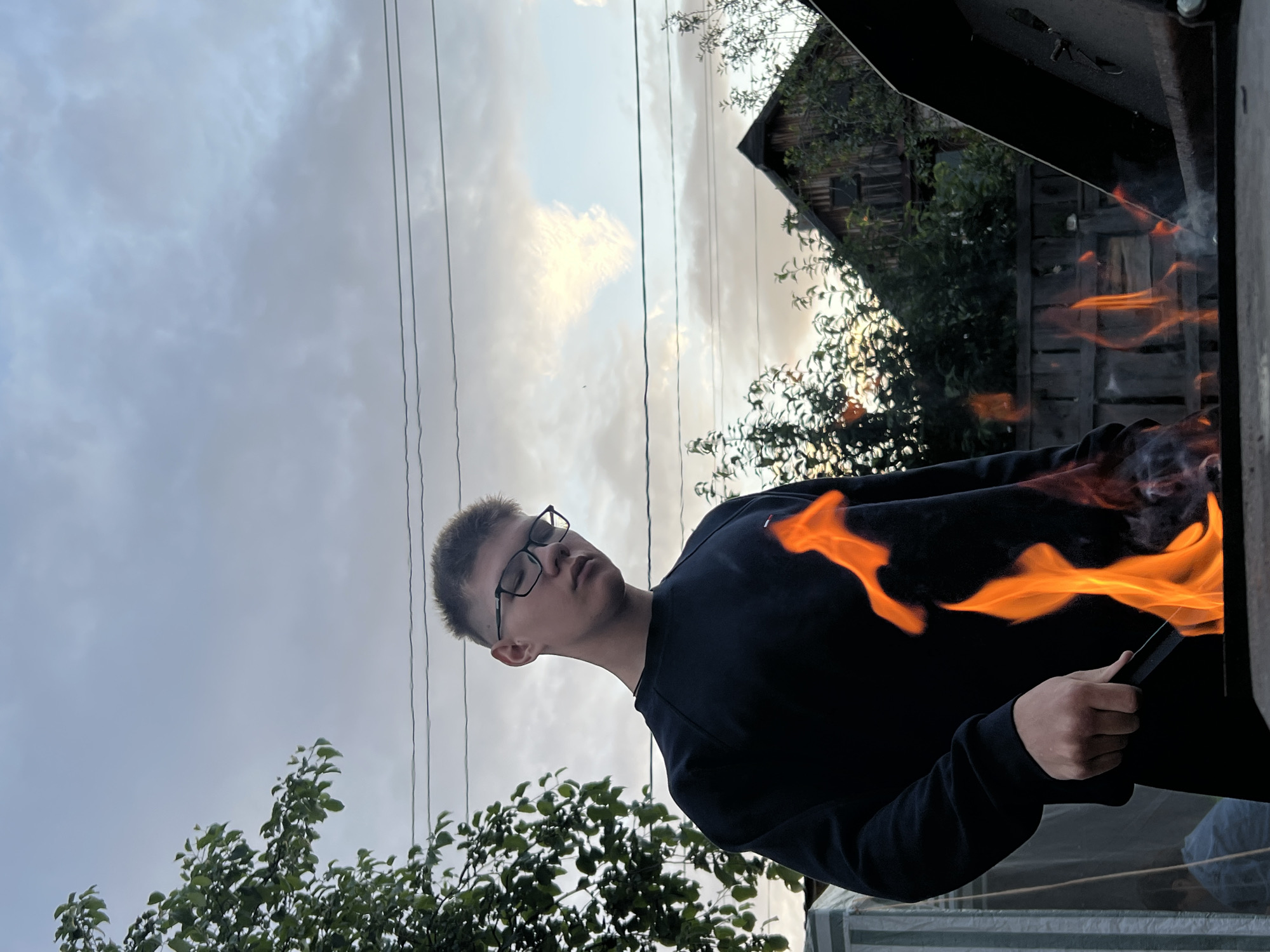 An eye-level, medium outdoor shot captures a young person with short brown hair and glasses standing in front of a barbecue with flames rising. The person is wearing a dark long-sleeved shirt and dark pants, and is holding tongs to tend to the fire. The background features a wooden fence, lush green trees and bushes, and a rustic wooden house. Above, a cloudy sky with hints of sunset colors looms, with power lines crossing the frame. The overall scene suggests a casual outdoor activity. The location is the city of Chaikovsky, Russia.
