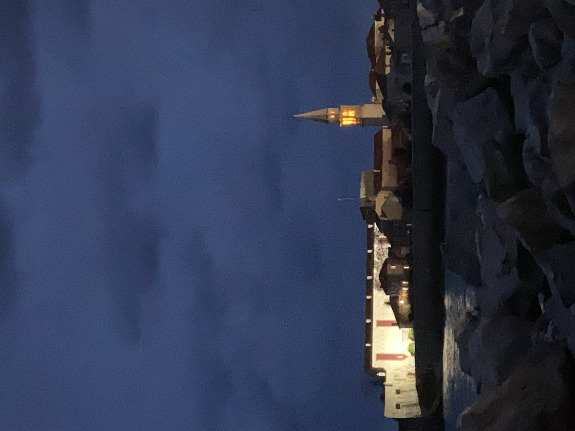 The image captures a night scene in what appears to be Budva, Montenegro, featuring a historic building with a distinct tower and a pointed roof, possibly a church or a bell tower, illuminated from within. The building has a light-colored facade with red accents and is bordered by a rocky, textured coastline on the right side of the frame. The sky is a dark blue, suggesting either twilight or night, and contrasts with the building's illuminated features. The image is shot at an angle, giving the scene a slightly disorienting perspective.