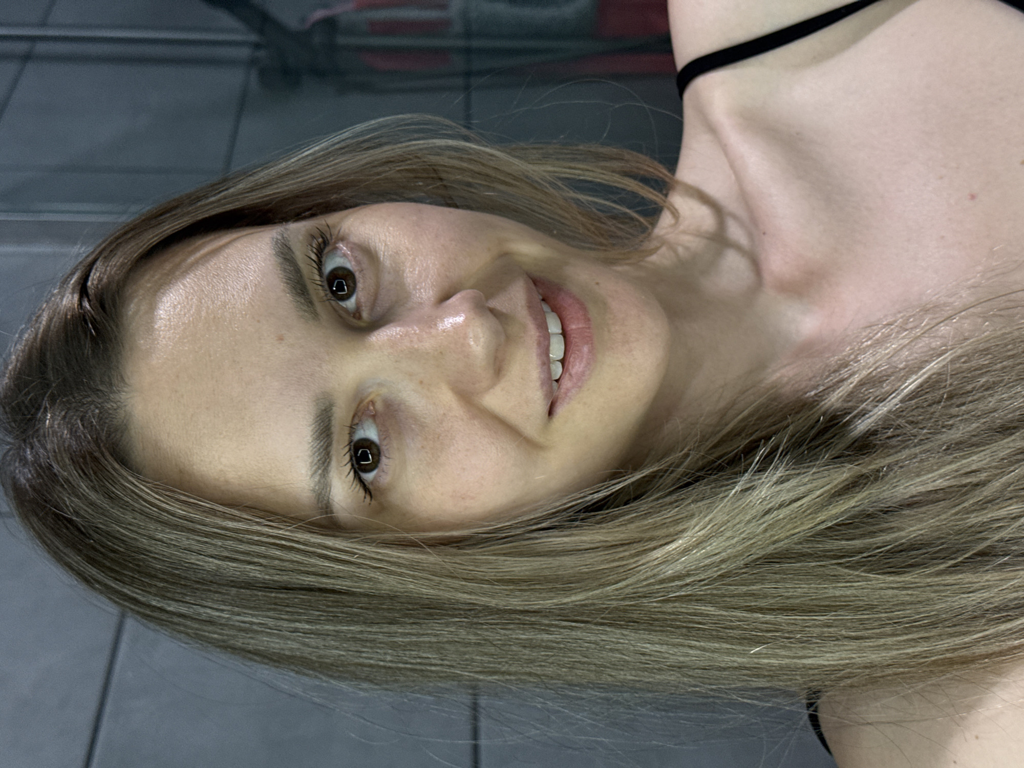 A close-up photograph shows a woman with light brown hair smiling. Her eyes are brown, and she has fair skin. She is wearing a black top. The background is a gray tiled wall with what appears to be a blurry reflection of exercise equipment. There is no evidence to establish that the image was taken in Chaikovsky, Russia.
