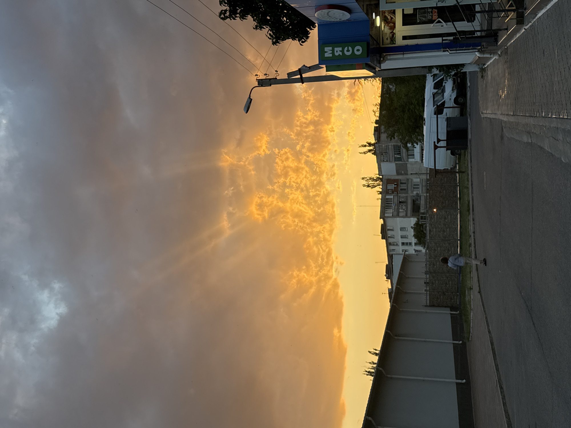 The image depicts a street scene in Tiraspol, Moldova, during a dramatic sunset. The sky is filled with clouds tinged in vibrant hues of orange and yellow, with rays of light beaming through them. In the foreground, a paved road runs diagonally across the frame. To the left, a long, light-colored building with a sloped roof dominates the scene, appearing to be a commercial or industrial structure. A person, dressed in a light-colored shirt and pants, is walking on the sidewalk in front of this building.
Across the street, on the right side of the image, a blue and green awning identifies a shop, with the word "МЯСО" (meat in Russian) visible on its signage. A street lamp stands tall next to the shop. Behind the shop, trees and several white vans are parked. In the background, apartment buildings with multiple stories are visible, suggesting an urban or semi-urban environment. The overall impression is a moment captured at dusk in a city setting, characterized by striking natural light and everyday urban elements.