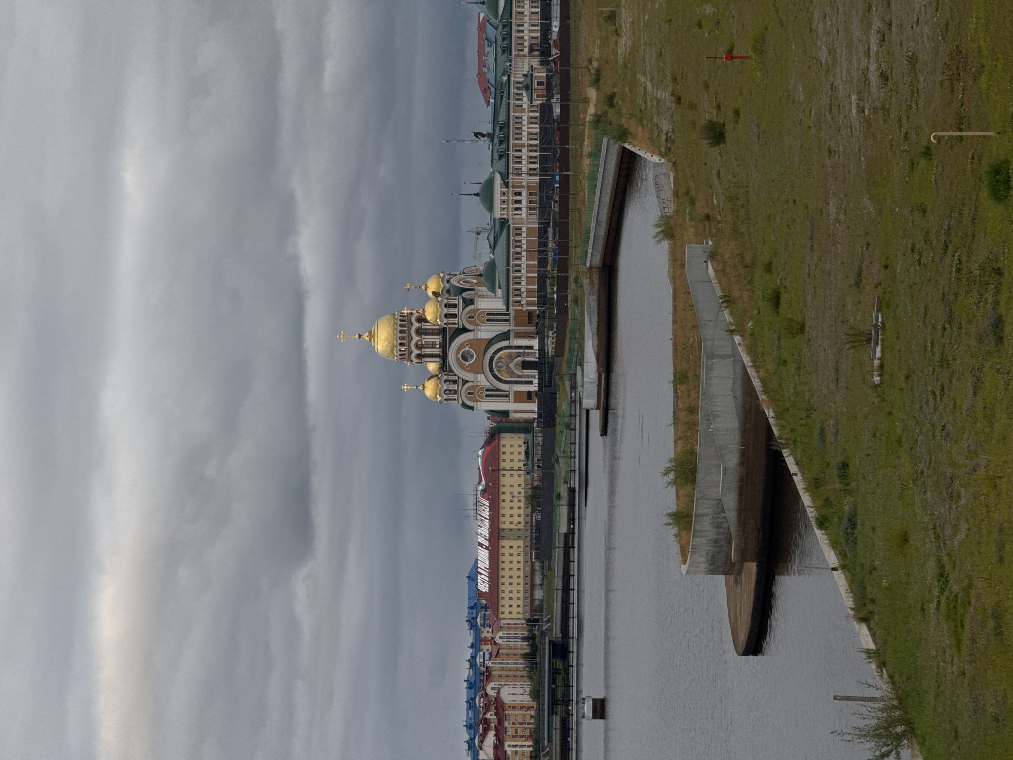 The image depicts the Holy Assumption Cathedral in Salekhard, Russia, a prominent landmark situated on the banks of the Poluy River. The cathedral, with its distinctive golden domes, stands against a dramatic backdrop of cloudy, overcast skies. In the foreground, a section of the riverbank is visible, featuring a curved concrete structure and grassy terrain. Beyond the cathedral, a line of buildings forms the city's skyline, with a bridge extending across the river. The overall scene conveys a sense of solemnity and architectural grandeur within an urban environment.
