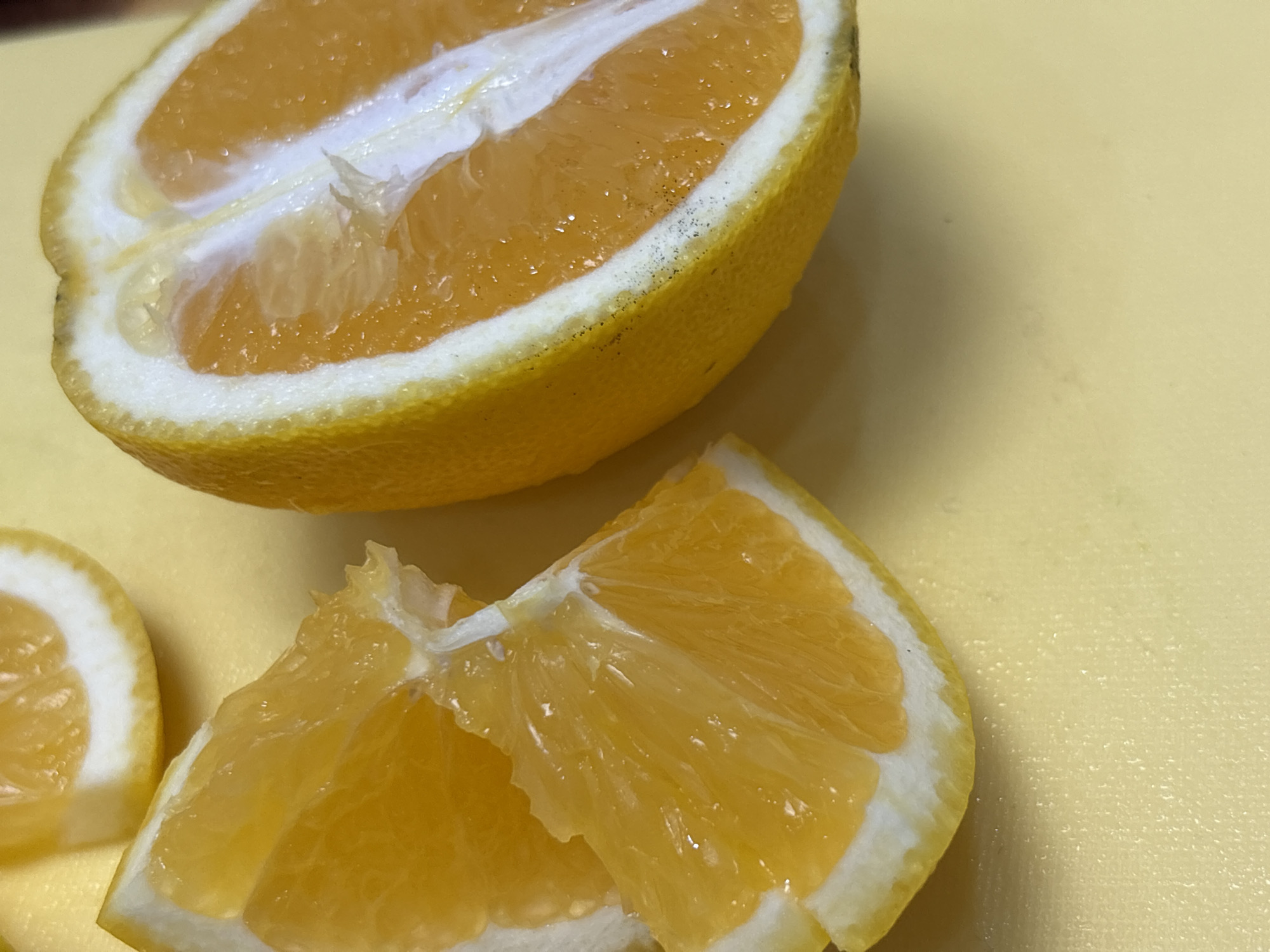 The image shows a close-up view of cut oranges on a pale yellow surface. On the left side of the frame is half of an orange, showing its segmented interior and thick rind. To the right, there are several wedges of orange, some overlapping, with their juicy flesh and white pith clearly visible. The lighting is bright, highlighting the vibrant colors of the fruit. There are no people, animals, or specific urban features visible that would indicate a location in Засечное, Russia.