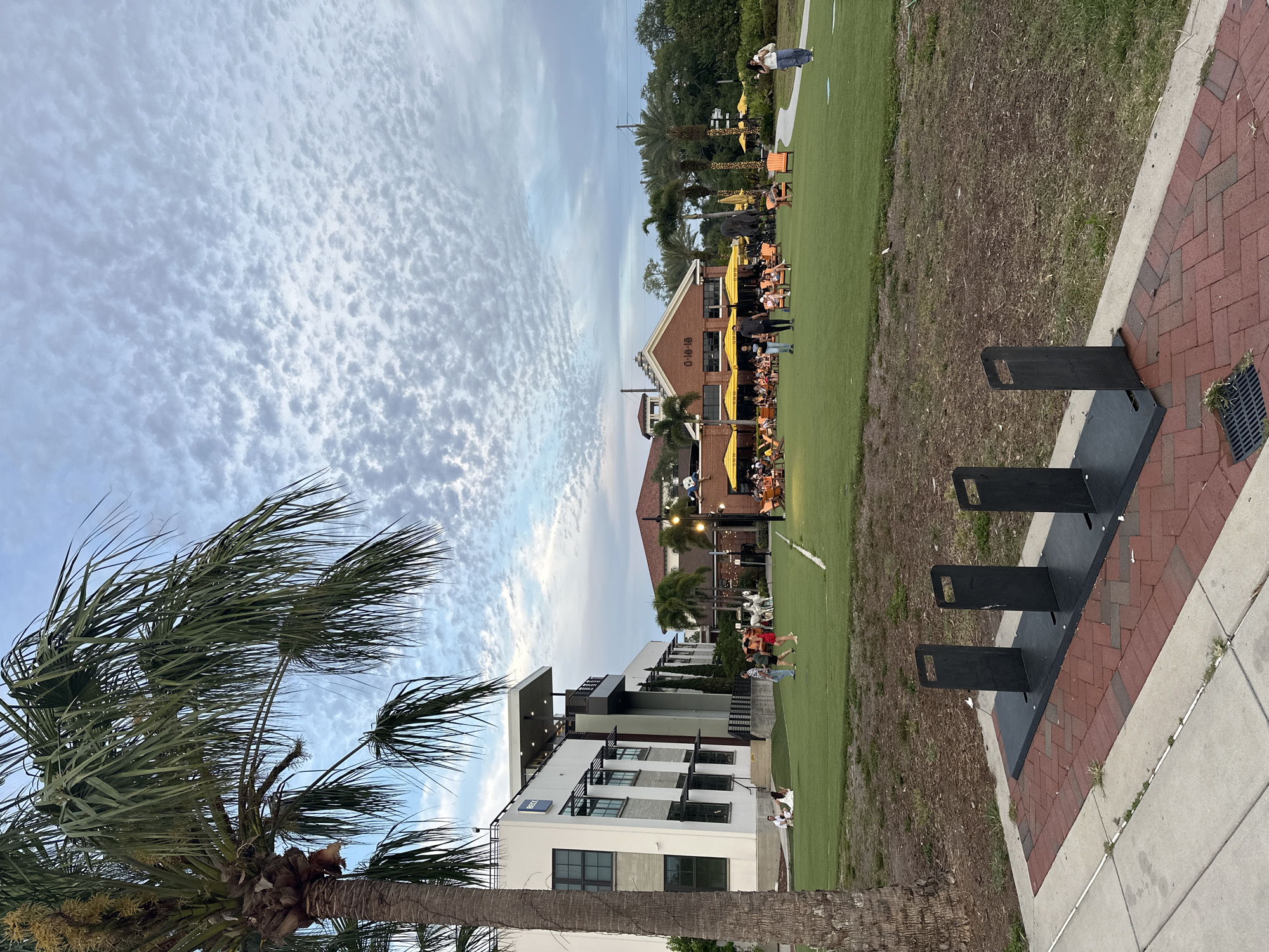 The image depicts an outdoor scene in Tampa, Florida, United States. The sky is partly cloudy. A tall palm tree is on the left side. A modern, multi-story building with large windows and a flat roof is located in the lower center. A red brick building with a peaked roof and yellow umbrellas shading outdoor seating is in the background. A grassy area is visible in front of these buildings, with people walking and seated at tables. In the foreground, there are three black metal structures, possibly bike racks, situated on a paved area with brick accents.