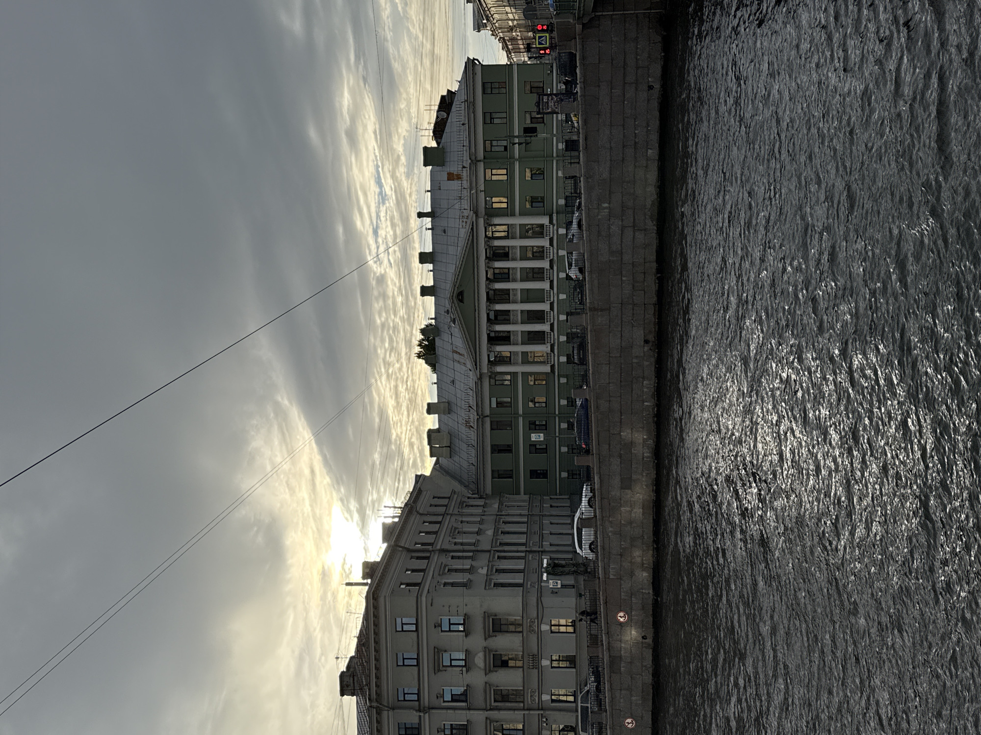 The image captures a wide-angle view of a canal in Saint Petersburg, Russia, under an overcast sky with the sun attempting to break through the clouds. On the left, a light-colored multi-story building with numerous windows and a mansard roof stands along the canal. Adjacent to it, to the right, is a larger, pale green building featuring a classical portico with white columns and a flat roof. Several cars are parked along the street in front of these buildings, and a few pedestrians are visible on the sidewalk. The canal itself is dark and choppy, reflecting the muted light from the sky. A stone embankment lines the canal in the foreground, with two circular signs visible on it. Overhead, several dark electrical wires crisscross the sky. The overall atmosphere is subdued, characteristic of a cloudy day in the city.