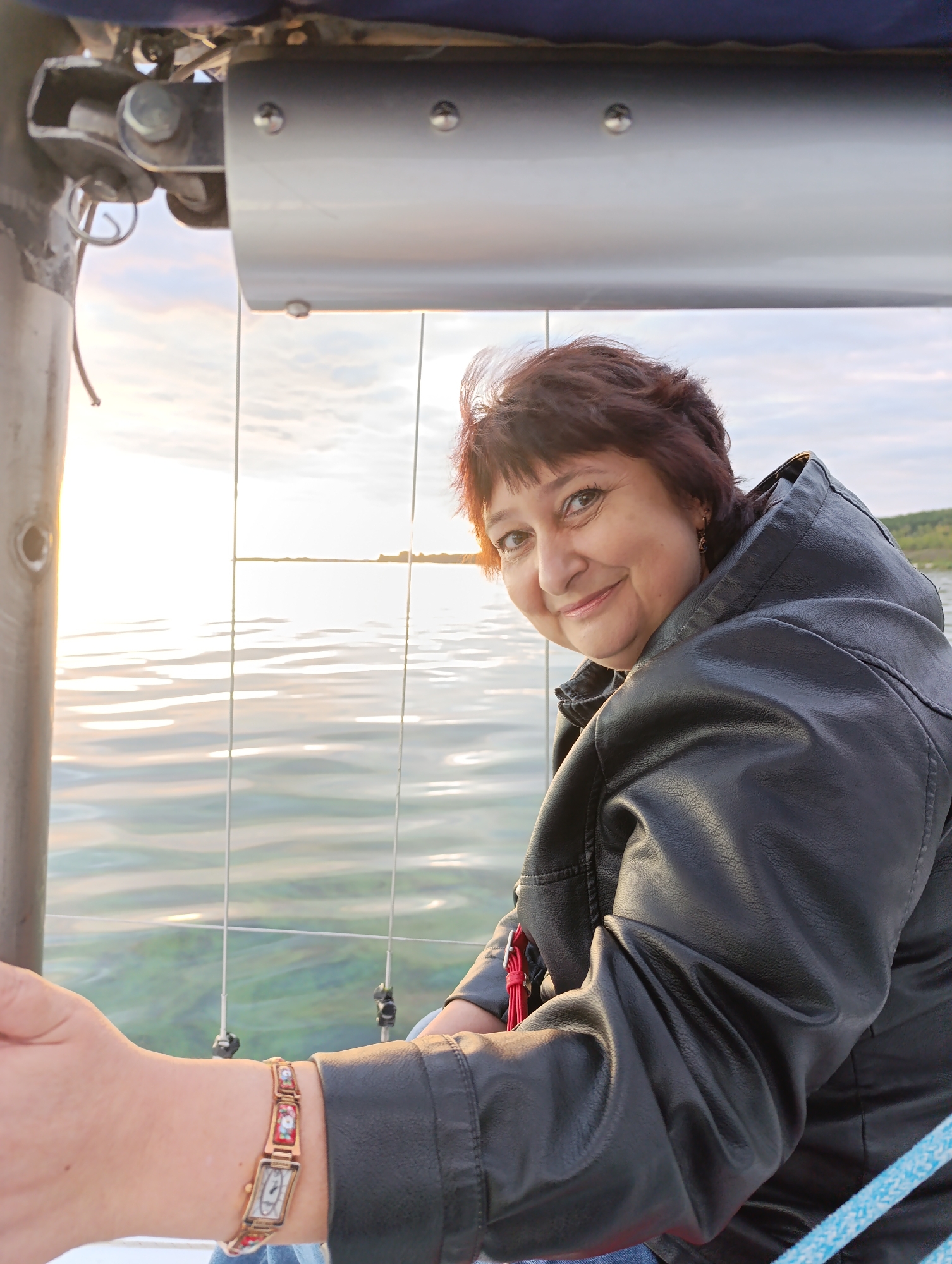 A woman with short, dark hair and a smiling expression is seated in a hot air balloon basket. She is wearing a black leather jacket and a bracelet with a patterned design. The basket has metal framework visible, and beyond it, a body of water reflects the sky. The sun is low in the sky, casting a warm glow on the scene. The background shows a hazy horizon with a hint of land. The specific location within Tolyatti, Russia, is not determinable from the image.