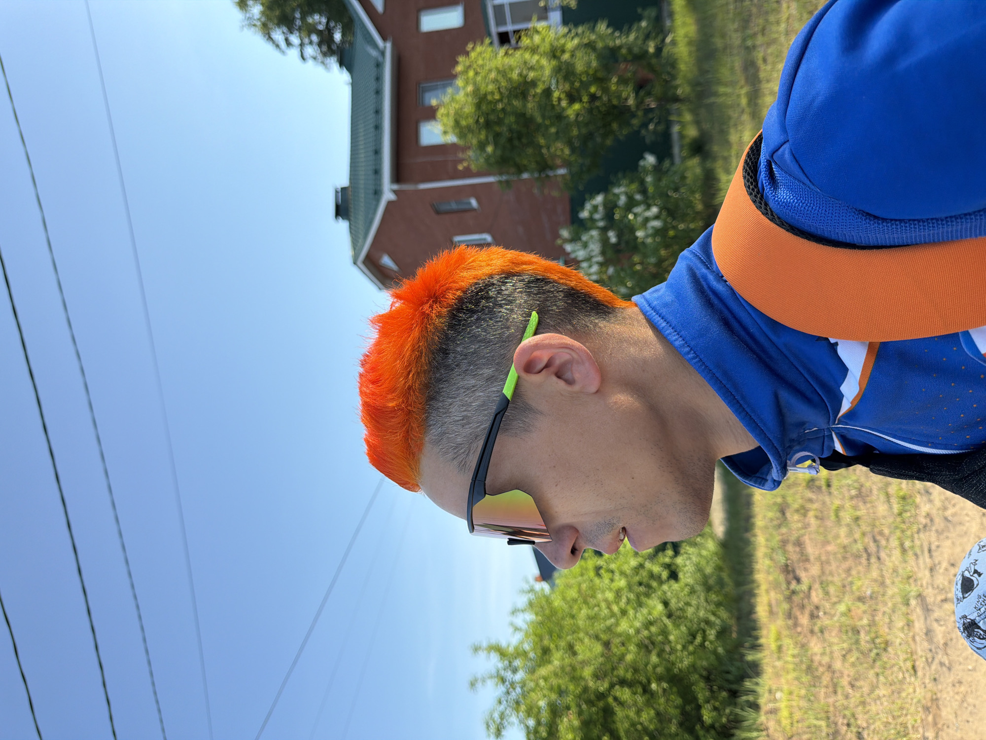 A medium-angle outdoor shot depicts a man with short, spiky orange hair and sunglasses with green accents, facing slightly right of frame. He wears a blue athletic jacket with an orange collar. In the background, there is a building with a red brick facade and a green roof, partially obscured by trees. Overhead, power lines crisscross the clear blue sky. The ground beneath the man appears to be grassy, with some bare patches. The location is Chaikovsky, Russia.