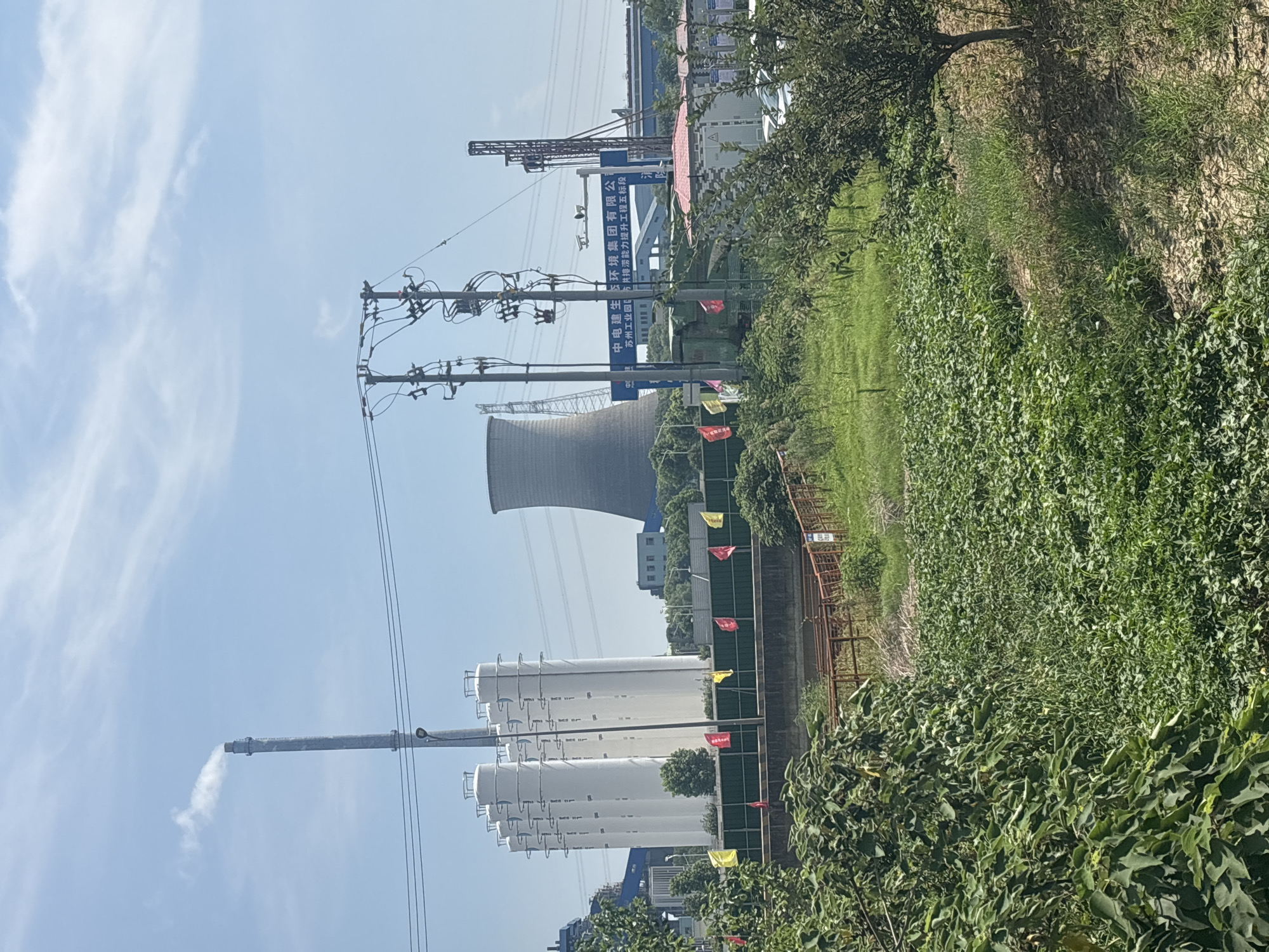 The image depicts a wide view of an industrial area in Suzhou Industrial Park, China, under a clear blue sky with scattered clouds. In the foreground, a lush green field of low-lying crops, possibly sweet potatoes, stretches across the lower half of the frame. Overgrown bushes and trees frame the left and right sides of the image, with a rusty metal fence running horizontally across the middle ground.
Behind the fence, several tall, white cylindrical silos stand in a row, appearing to be part of a larger industrial complex. To the right of the silos, a large, grey, hyperboloid cooling tower dominates the mid-ground. Smoke or steam is visible emerging from a tall industrial smokestack to the left of the silos, indicating ongoing industrial activity.
Further in the background, a blue sign with Chinese characters is mounted on a structure, likely identifying the facility. Electrical transmission towers and wires are visible throughout the scene, highlighting the industrial infrastructure. In the far background, more industrial buildings and structures can be seen, suggesting a sprawling manufacturing or power generation facility. The overall impression is one of a functional industrial zone with surrounding agricultural or undeveloped land.