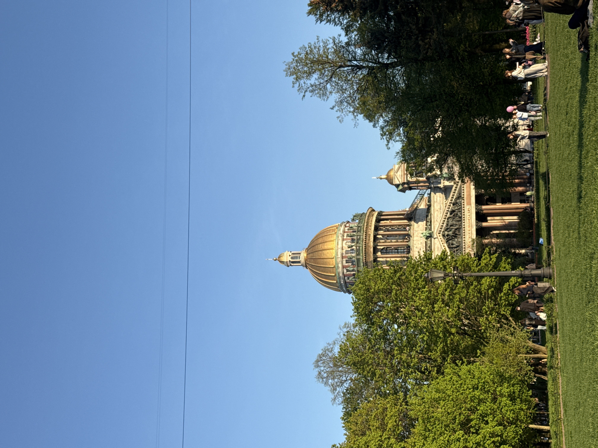 The image shows the dome of St. Isaac's Cathedral in Saint Petersburg, Russia, framed by trees. Clear blue sky fills the left side of the image. The cathedral's golden dome is centrally positioned. People are visible in the background, along the grass, near the trees. A lamppost stands amongst the trees.