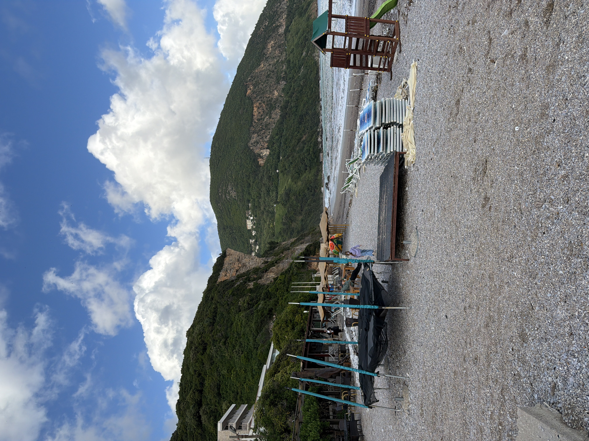 The image depicts a rocky beach in Prijevor, Montenegro, with a mountain as a backdrop. The sky is blue with scattered white clouds. On the beach, there are stacked beach chairs and a wooden playset. In the foreground, beach umbrellas are being prepared, with blue poles visible. There are buildings at the bottom left of the frame. The beach appears mostly empty. The sea is visible in the middle ground between the stacked chairs and the mountain.