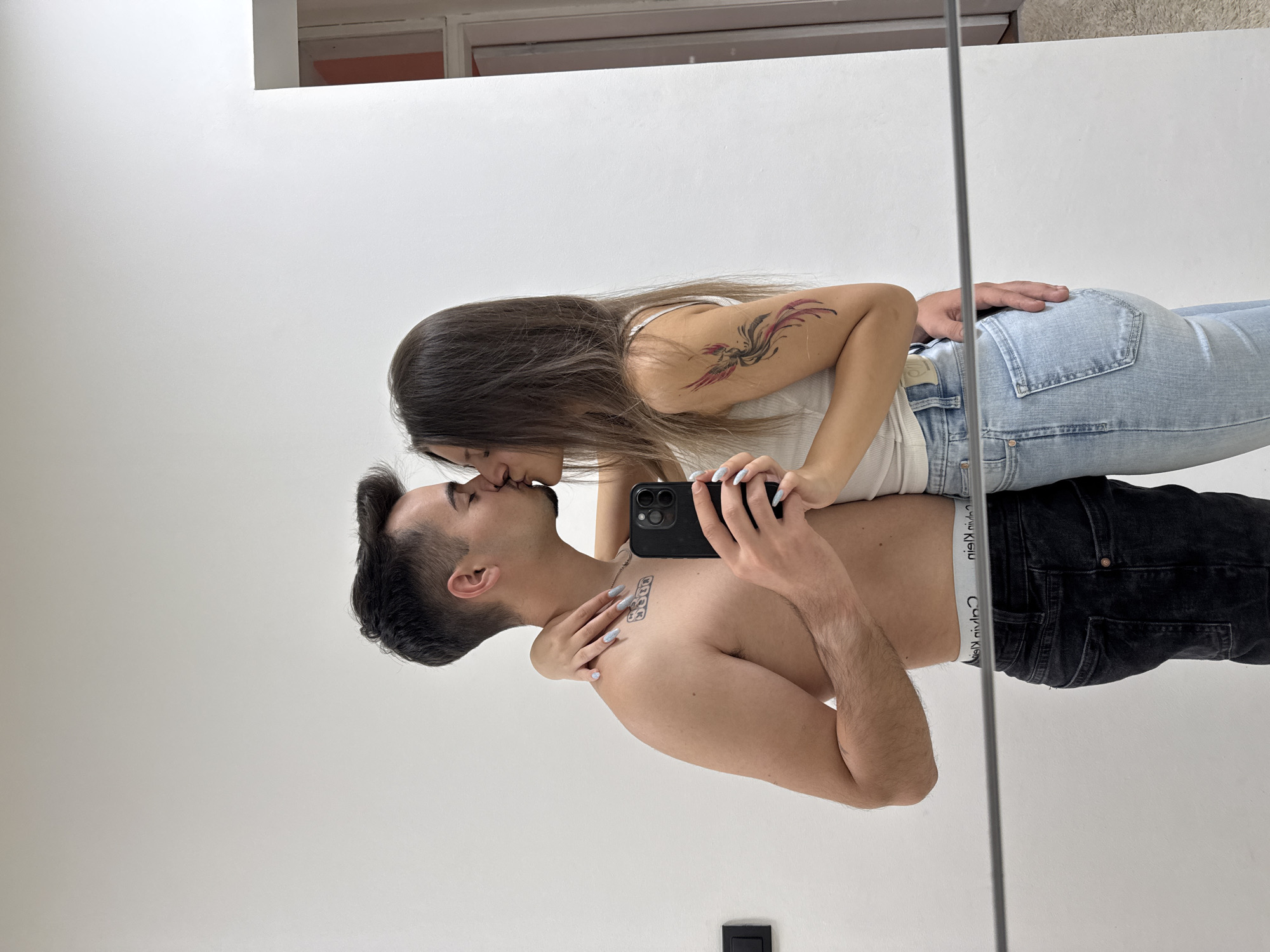 The image depicts two individuals, a male and a female, kissing each other while taking a mirror selfie in an indoor setting in Sofia, Bulgaria. The male, positioned on the left, is shirtless, exposing a tattoo reading "5800" on his upper left chest/shoulder area. He has short, dark hair and light stubble. He is wearing dark wash jeans with the waistband of Calvin Klein underwear visible above. He holds a black smartphone with a multi-lens camera array in his right hand, positioned in front of his torso, capturing the reflection. His left arm is around the female's waist. The female, on the right, has long, straight dark hair and is wearing a white ribbed tank top and light wash blue jeans. A multi-colored tattoo, appearing to be a phoenix or similar bird design with red/pink and black elements, is visible on her upper left arm. Her right arm is wrapped around the male's neck, and her left hand rests on his shoulder. Both individuals exhibit long fingernails painted with light blue polish. The background consists of a plain white wall. A black electrical outlet or switch is visible on the wall to the left of the male. In the upper right, a doorway or opening with a white frame and a glimpse of a reddish-orange interior is visible. A dark, thin horizontal bar, possibly part of the mirror frame or room architecture, runs across the lower portion of the image. A light-colored carpet or rug is visible on the floor below the bar on the right side.