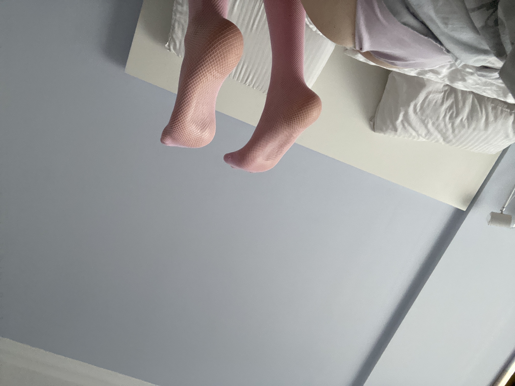 The image shows a close-up view of a person's legs, from the knees down, wearing pink fishnet stockings. The person appears to be sitting or lying on a bed, with pillows and blankets visible in the background. The setting seems to be a bedroom, with a light-colored headboard and a pale blue wall. The lighting suggests it might be daytime, possibly indoors. The perspective is unusual, as if the photo was taken from below, looking up at the legs. There is no visible text in the image. The overall mood is somewhat casual and relaxed, focusing on the texture and color of the stockings and the soft fabrics of the bedding.