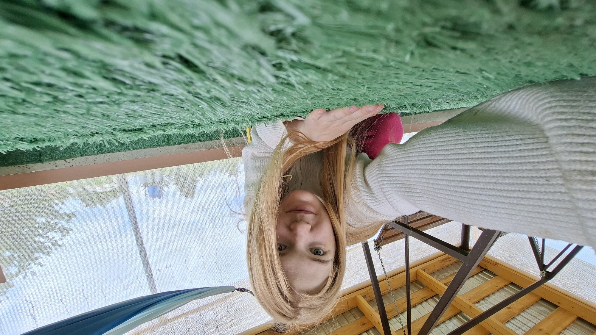 A young woman with blonde hair is captured in a low-angle, close-up selfie. She is wearing a white knitted sweater and pink pants. Her right hand is extended forward, resting on a green artificial turf surface, suggesting she is either in a garden, playground, or similar outdoor recreational area. The background reveals a wooden roof structure with metal beams, and a mesh screen that offers a view of trees and a blue water tower against an overcast sky. The setting appears to be outdoors, possibly in a rural or suburban location like Большой Букор, Russia. The overall scene is informal and personal, focusing on the woman's engagement with the camera and her surroundings.