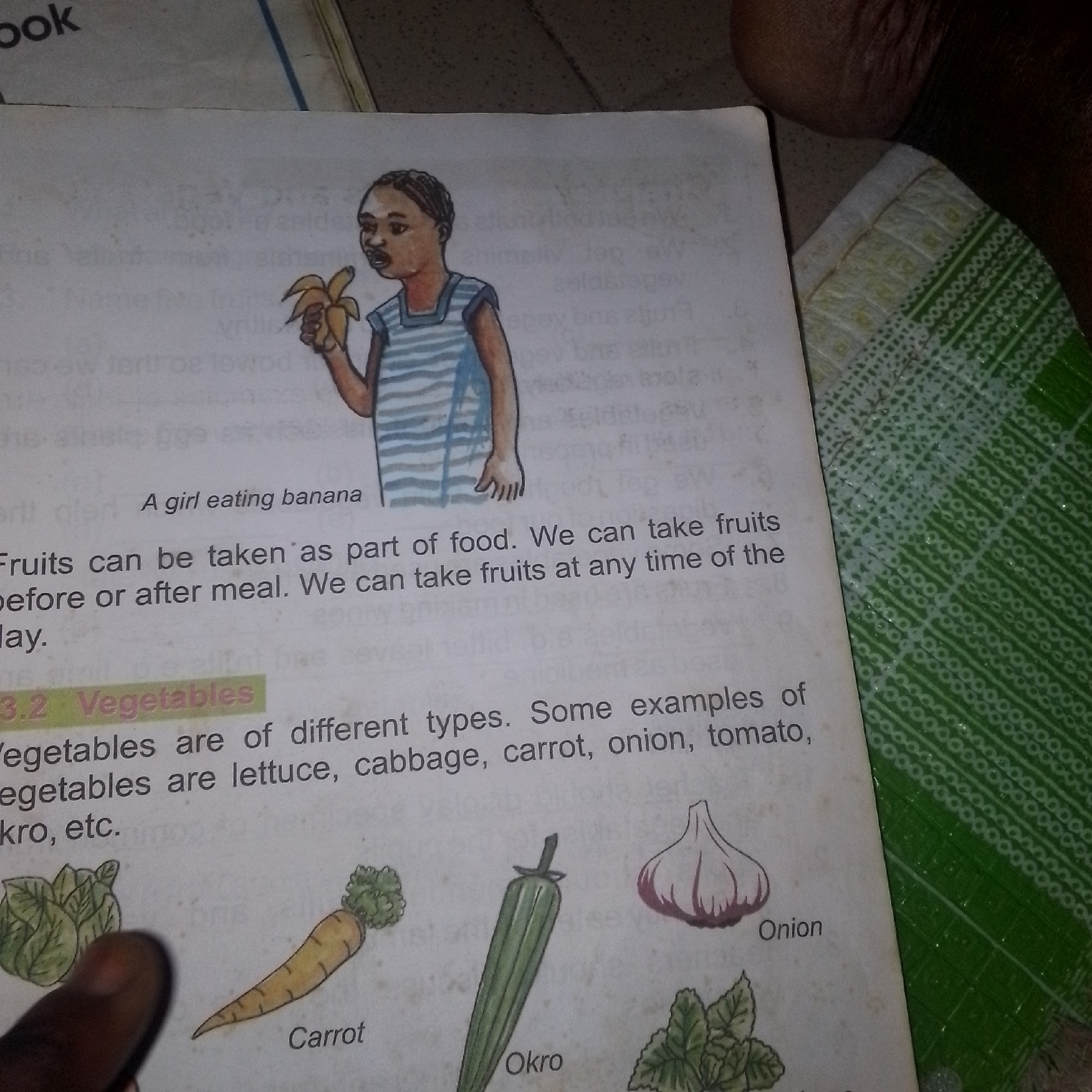 The image is an overhead view of an open book, likely a children's educational book, placed on a textured surface. The left page displays a cartoon illustration of a young girl eating a banana, with the caption "A girl eating banana" below it. Adjacent to the illustration, text discusses fruits: "Fruits can be taken as part of food. We can take fruits before or after meal. We can take fruits at any time of the day."
The right page, more prominently featured, has a section heading "3.2 Vegetables". Below this heading, text reads: "Vegetables are of different types. Some examples of vegetables are lettuce, cabbage, carrot, onion, tomato, Okro, etc." This text is accompanied by colorful illustrations of various vegetables, including cabbage, a carrot, an okra pod, and an onion. The book is positioned on a patterned green and white fabric, possibly a tablecloth or mat. A person's finger is visible at the bottom left, pointing towards the illustration of the cabbage. The background is indistinct, but appears to be a floor or ground surface.